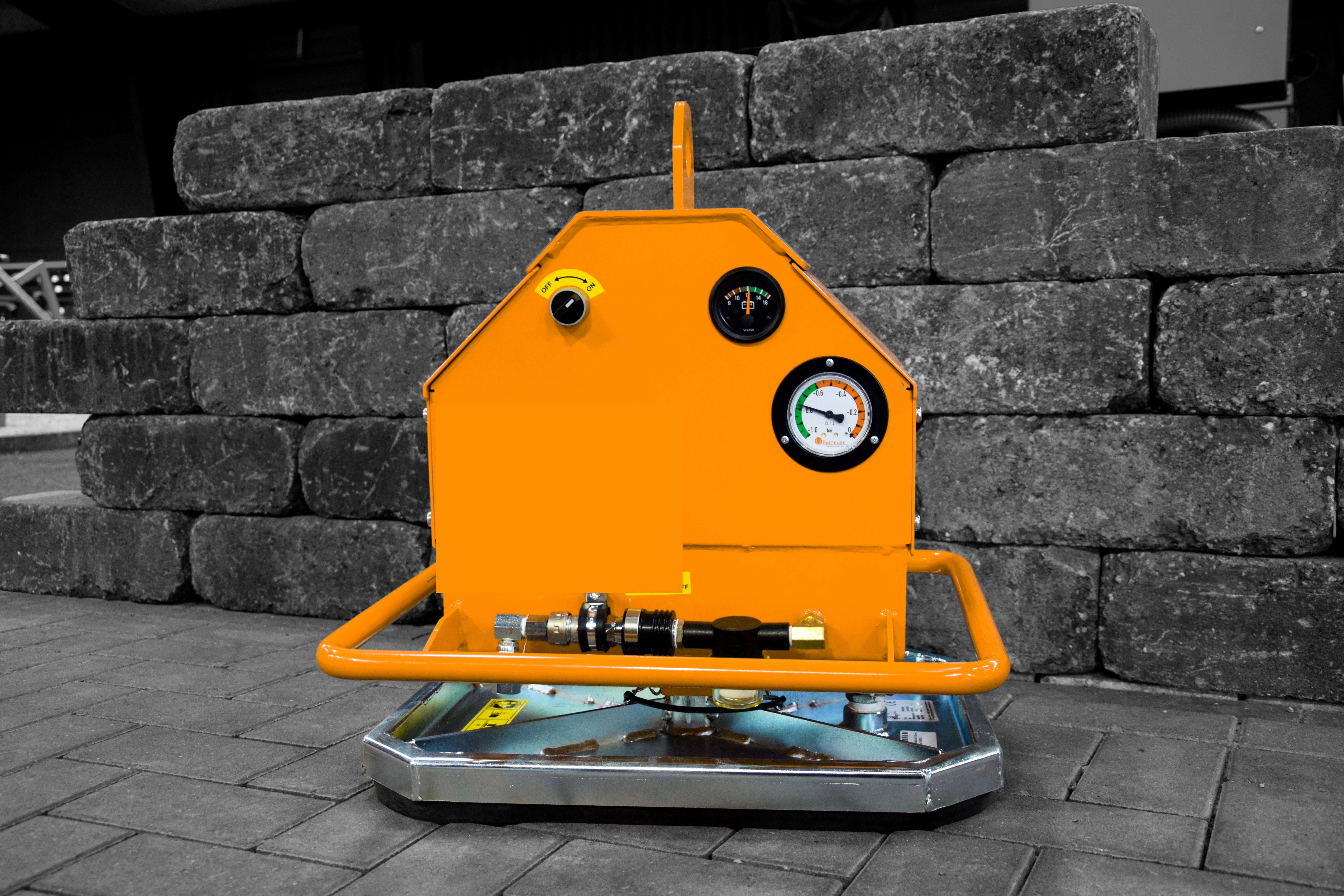 Ein orangefarbenes Vakuumhebegerät mit Messgeräten und Metallsockel steht auf einer gepflasterten Fläche vor Steinblöcken – Optimas.