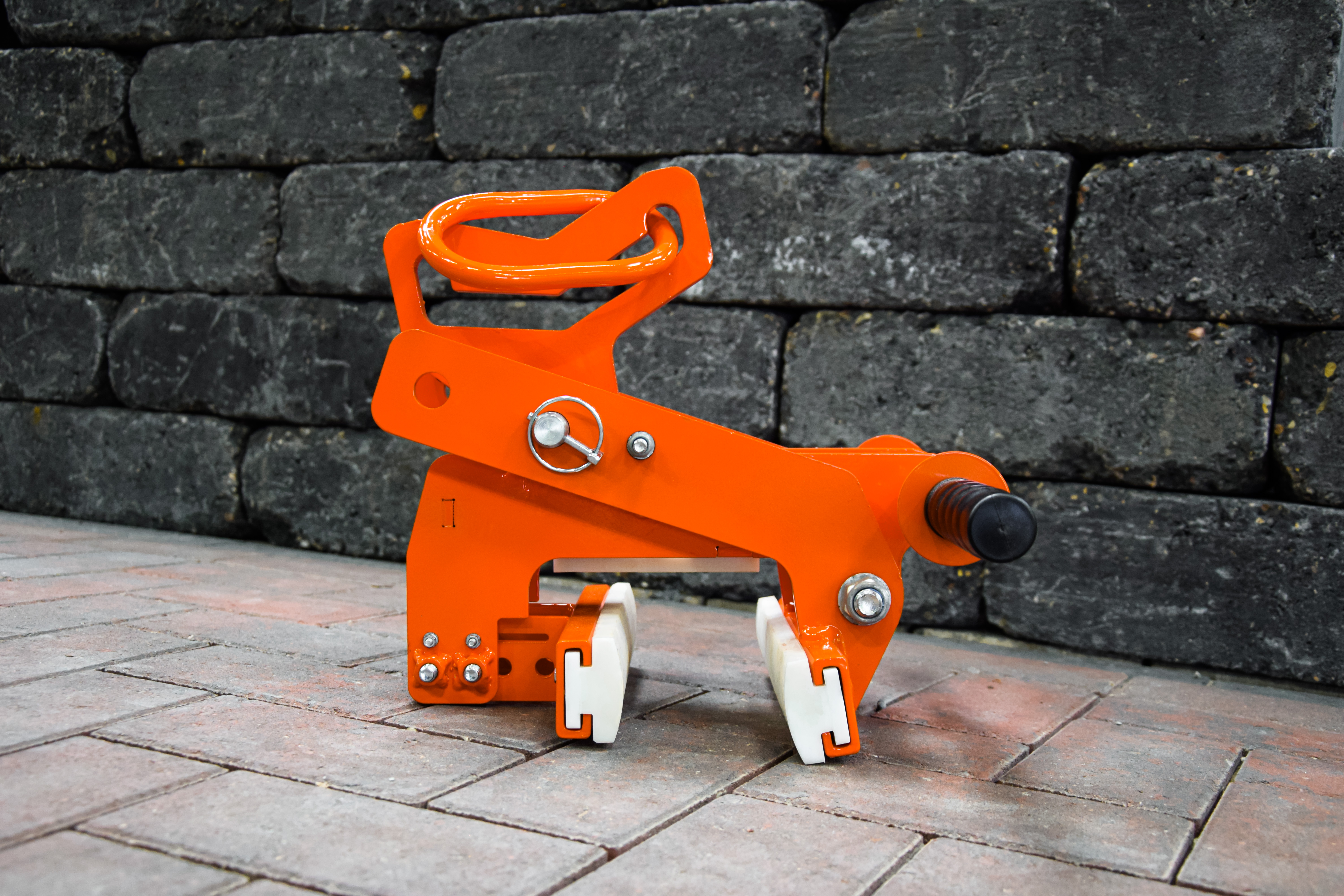 Eine orangefarbene mechanische Steinhebeklemme mit weißen Gummipads, die auf einer gepflasterten Fläche vor einer Wand steht – Optimas.