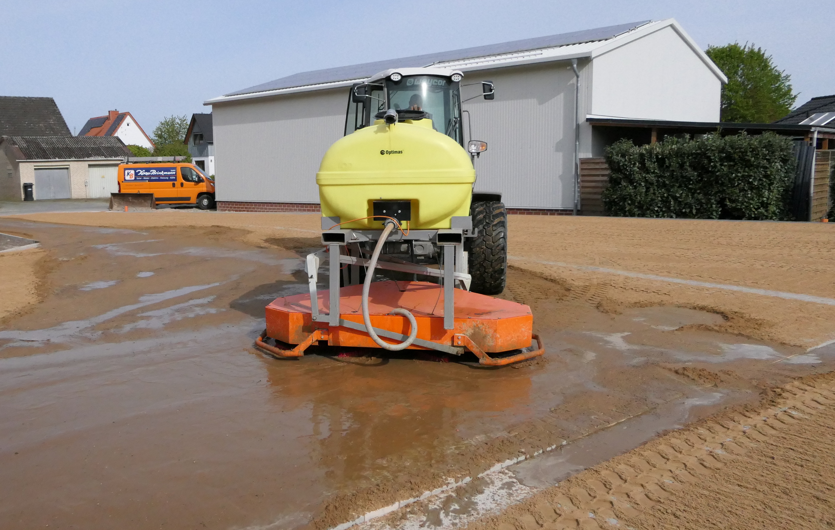 Eine gelbe Landmaschine mit großem Tank arbeitet auf einer sandigen Oberfläche und versprüht Wasser – Optimas.