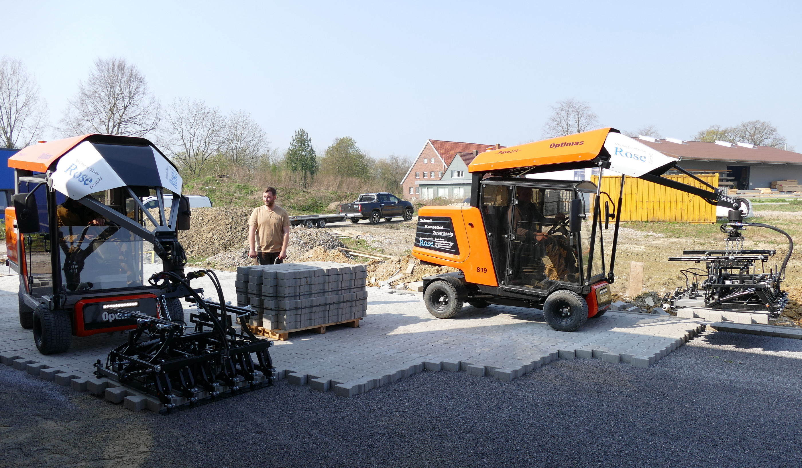 Zwei Optimas-Pflastermaschinen auf einer Baustelle, ein Mann, der neben einem Stapel Pflastersteine ​​steht, und ein LKW mit Anhänger im Hintergrund – Optimas