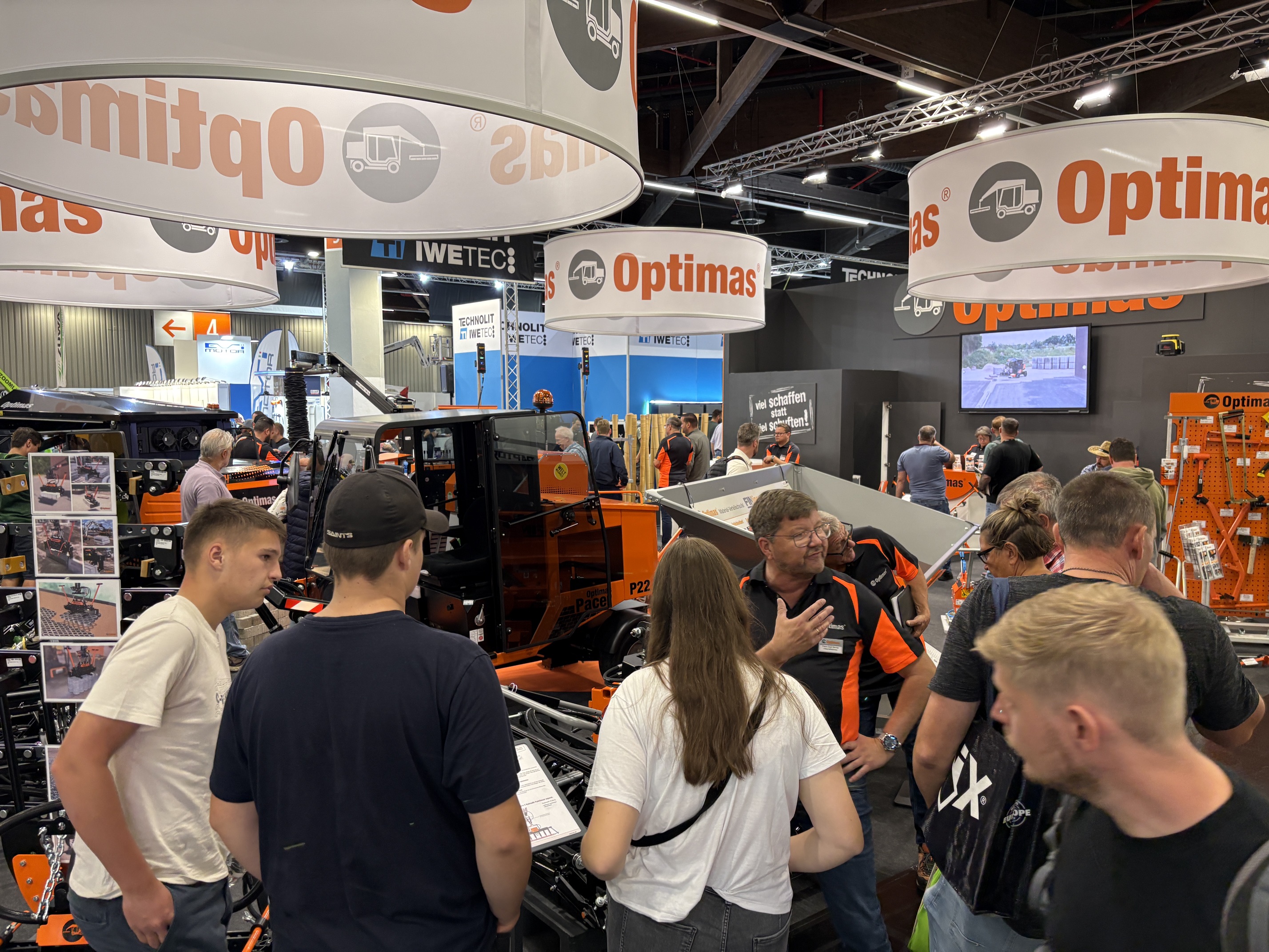 Besucher an einem Messestand betrachten Baumaschinen unter großen Hängeschildern der Marke Optimas – Optimas.