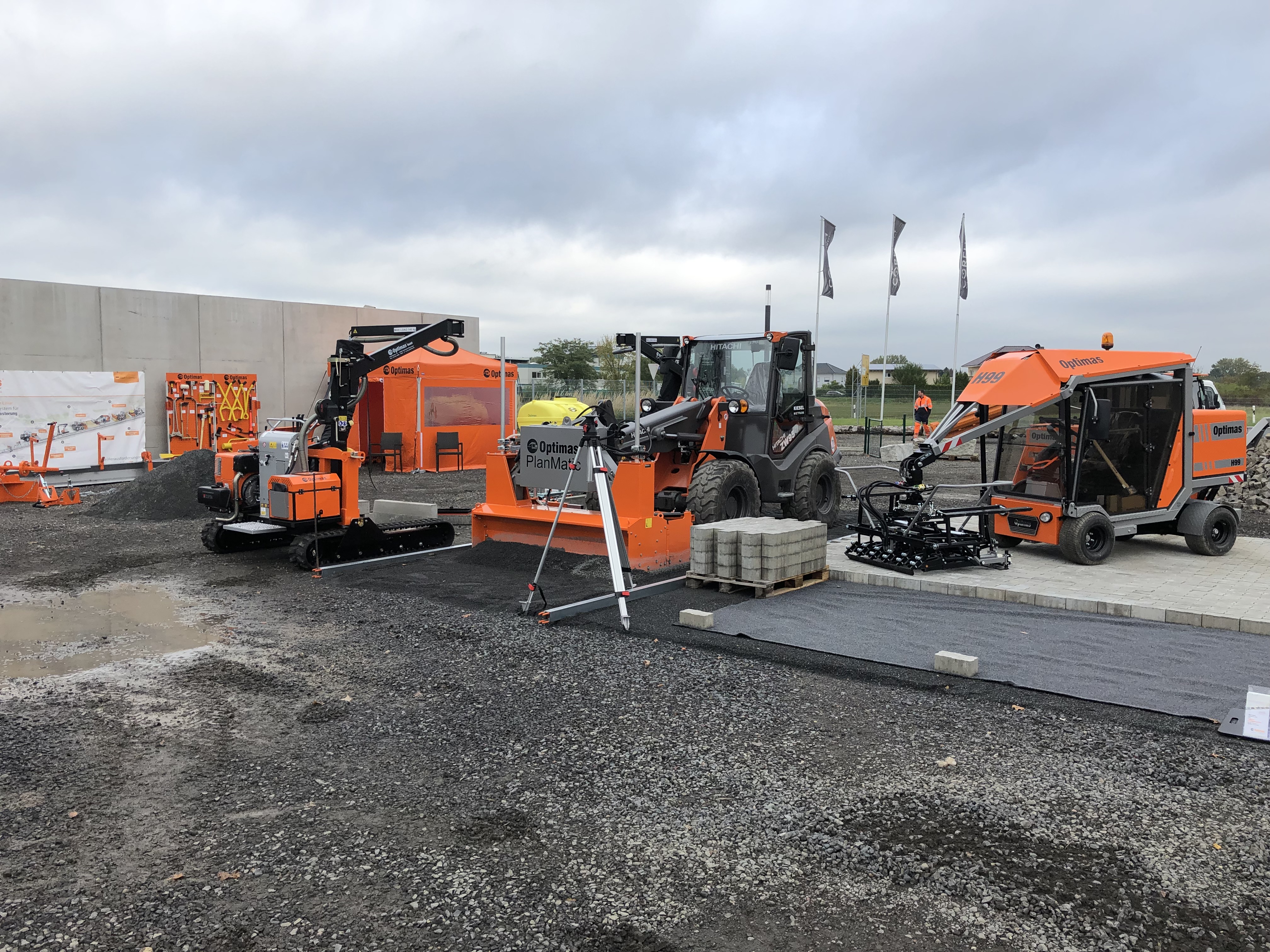 Während einer Ausstellung im Freien werden verschiedene orangefarbene Optimas-Baufahrzeuge auf einer Kiesfläche ausgestellt, wodurch ein auffälliges Display entsteht – Optimas.