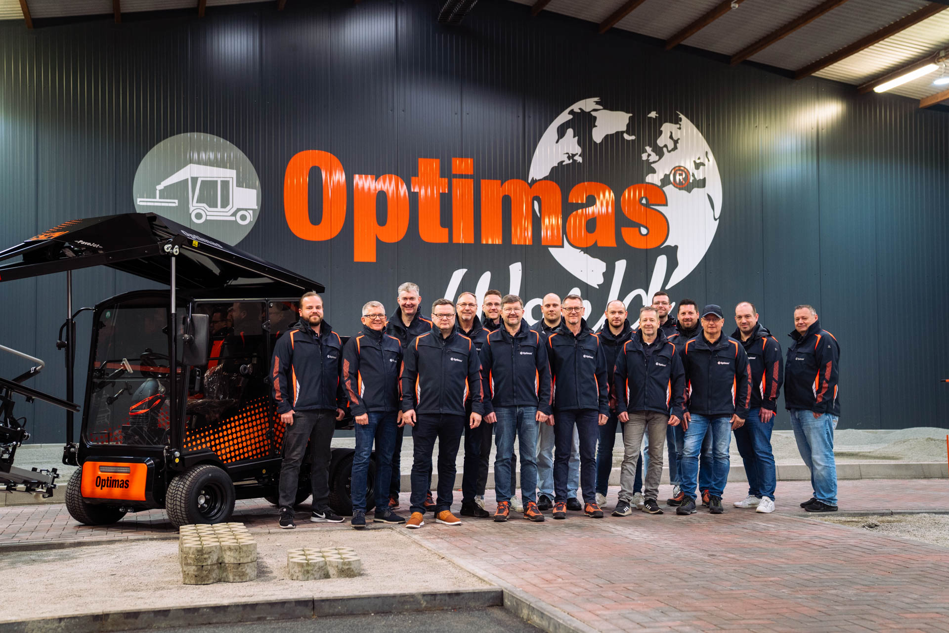 A group of employees in jackets stands in front of the Optimas logo and a vehicle, showcasing teamwork – Optimas.