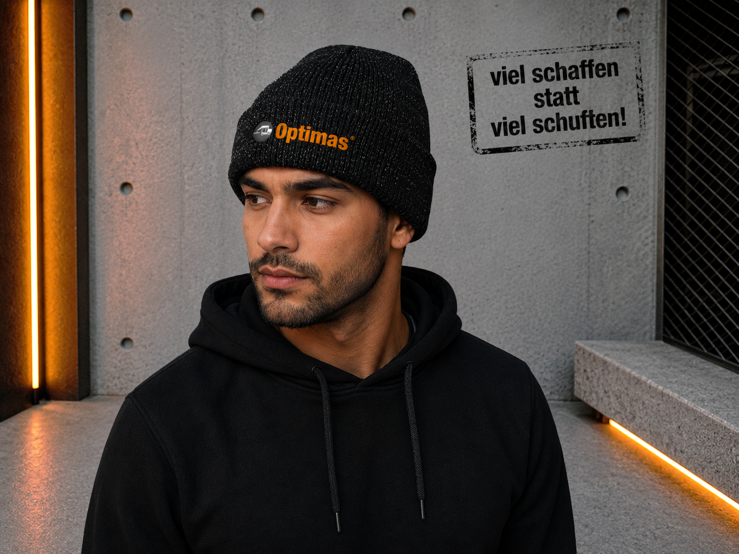 Ein Mann mit schwarzer Mütze und Kapuzenpullover posiert vor einer strukturierten Wand mit orangefarbener Beleuchtung – Optimas.