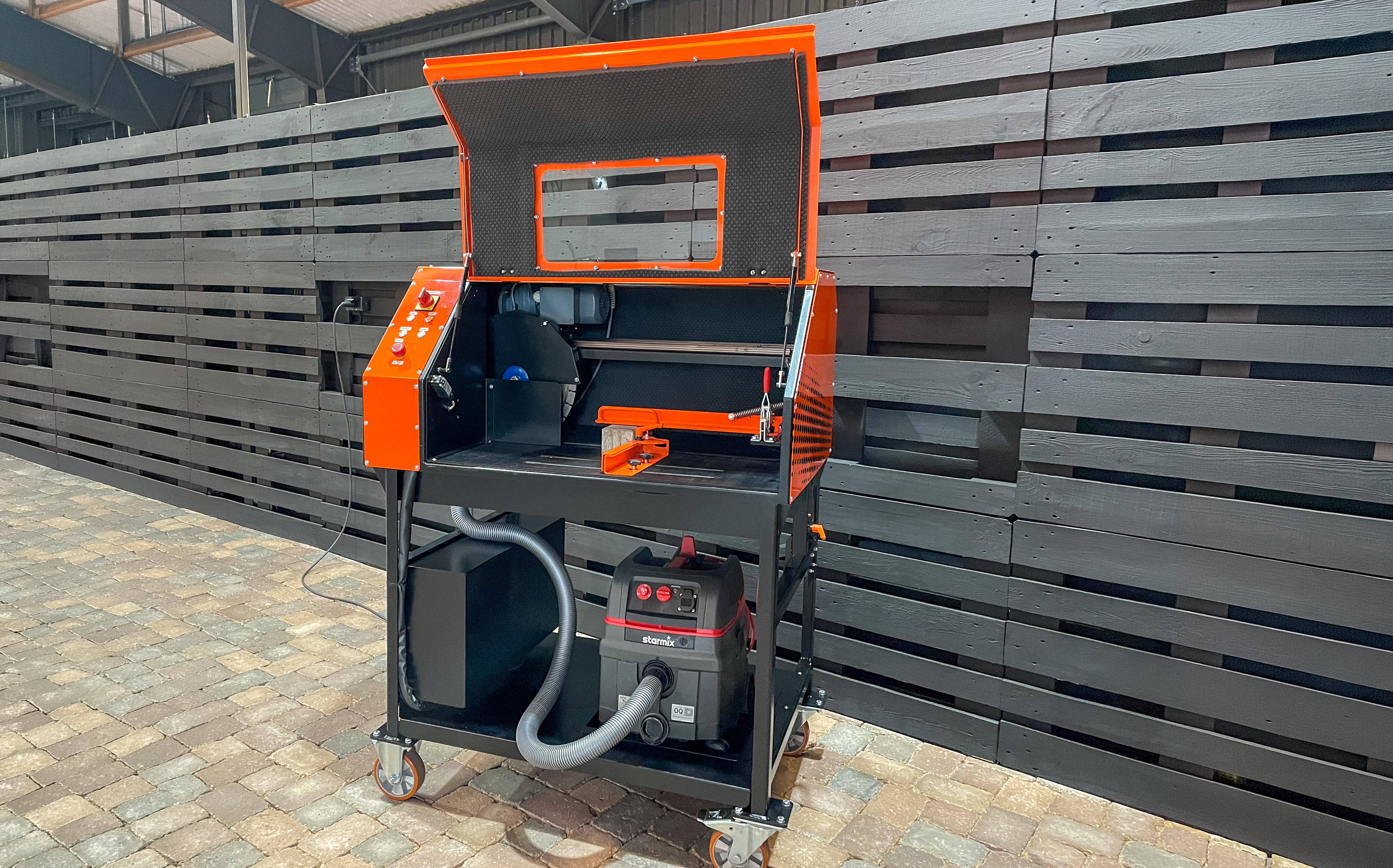 Eine schwarz-orangefarbene Holzbearbeitungsmaschine mit Bedienelementen und Staubabscheider vor einer Holzwand – Optimas.