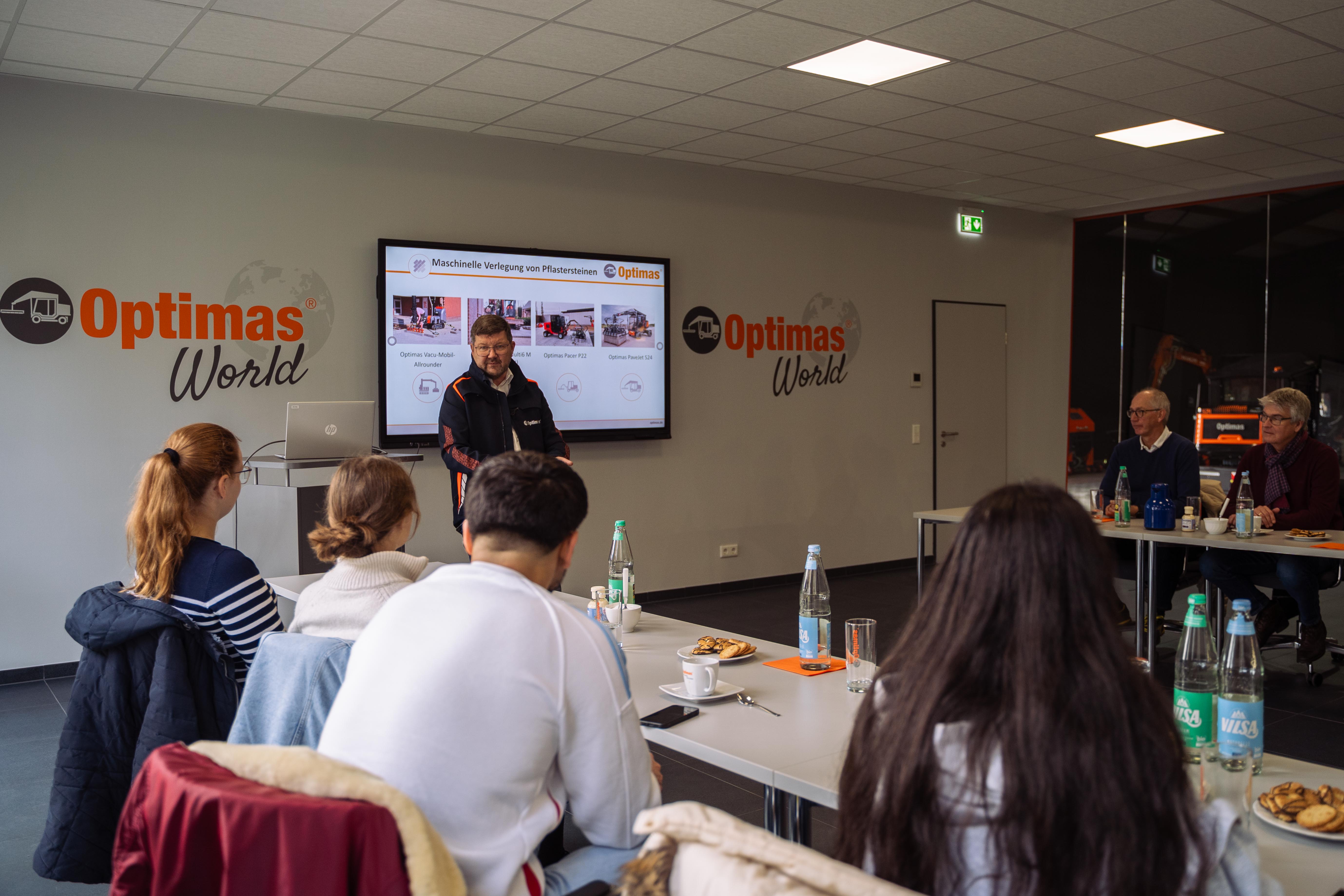 Eine Präsentation in der Optimas World, bei der die Teilnehmer an Tischen sitzen und mit einem Redner und visuellen Hilfsmitteln interagieren – Optimas.