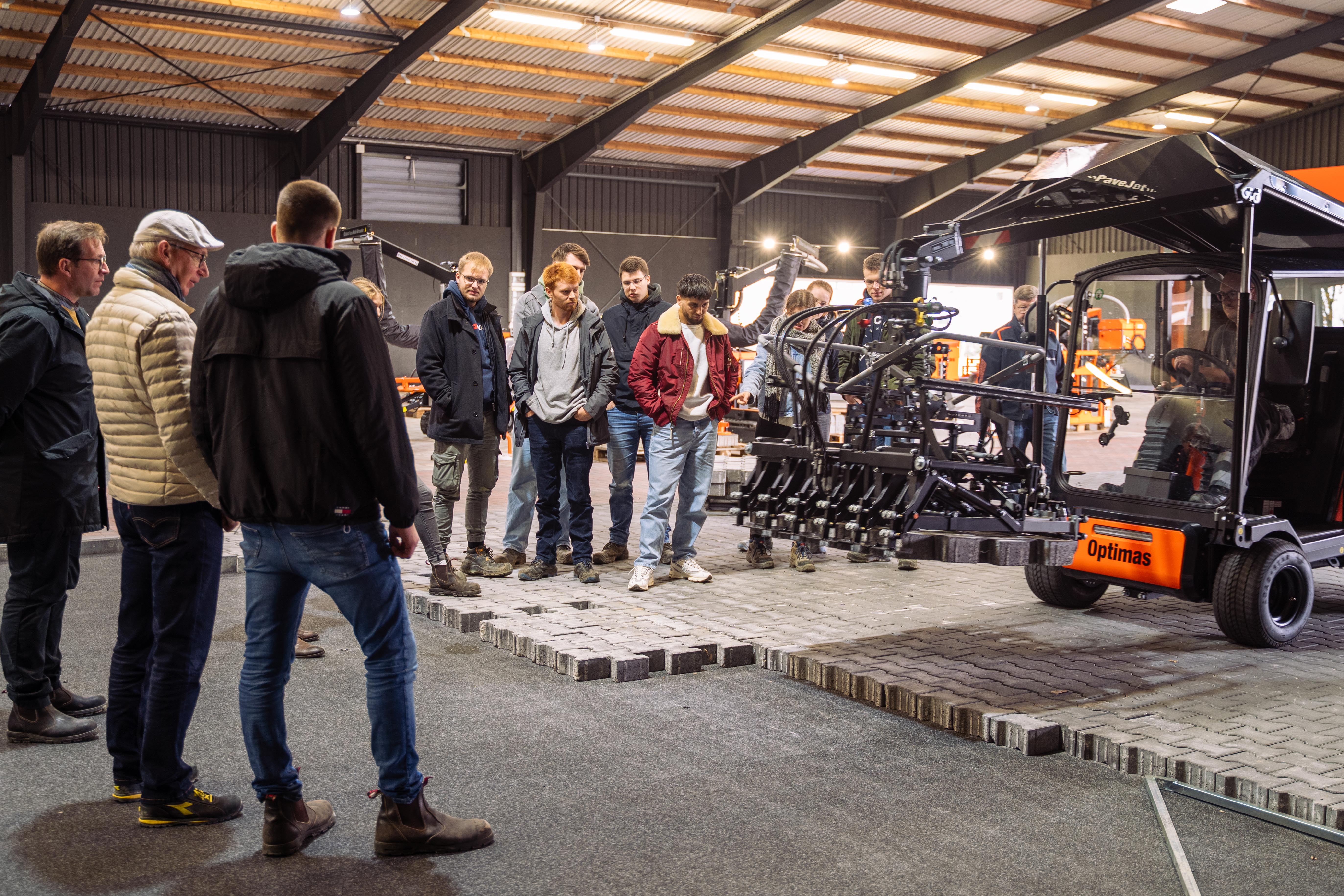 Eine Gruppe von Menschen beobachtet eine Maschinenvorführung in einem großzügigen Innenbereich – Optimas.