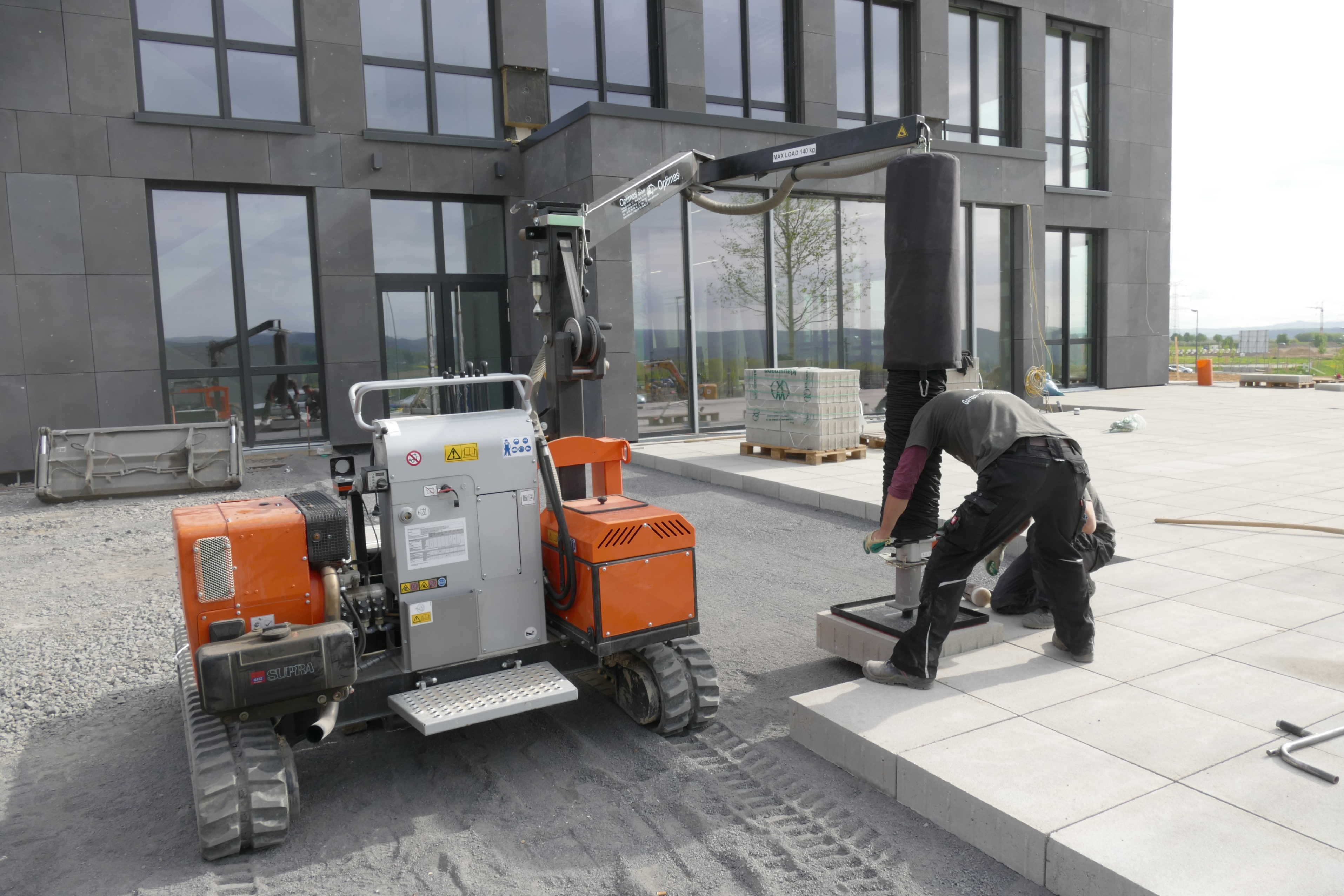 Eine Baustelle zeigt Arbeiter, die einen Minikran verwenden, um Betonpflastersteine ​​in der Nähe eines modernen Gebäudes zu installieren, wobei Effizienz und Präzision im Vordergrund stehen – Optimas.