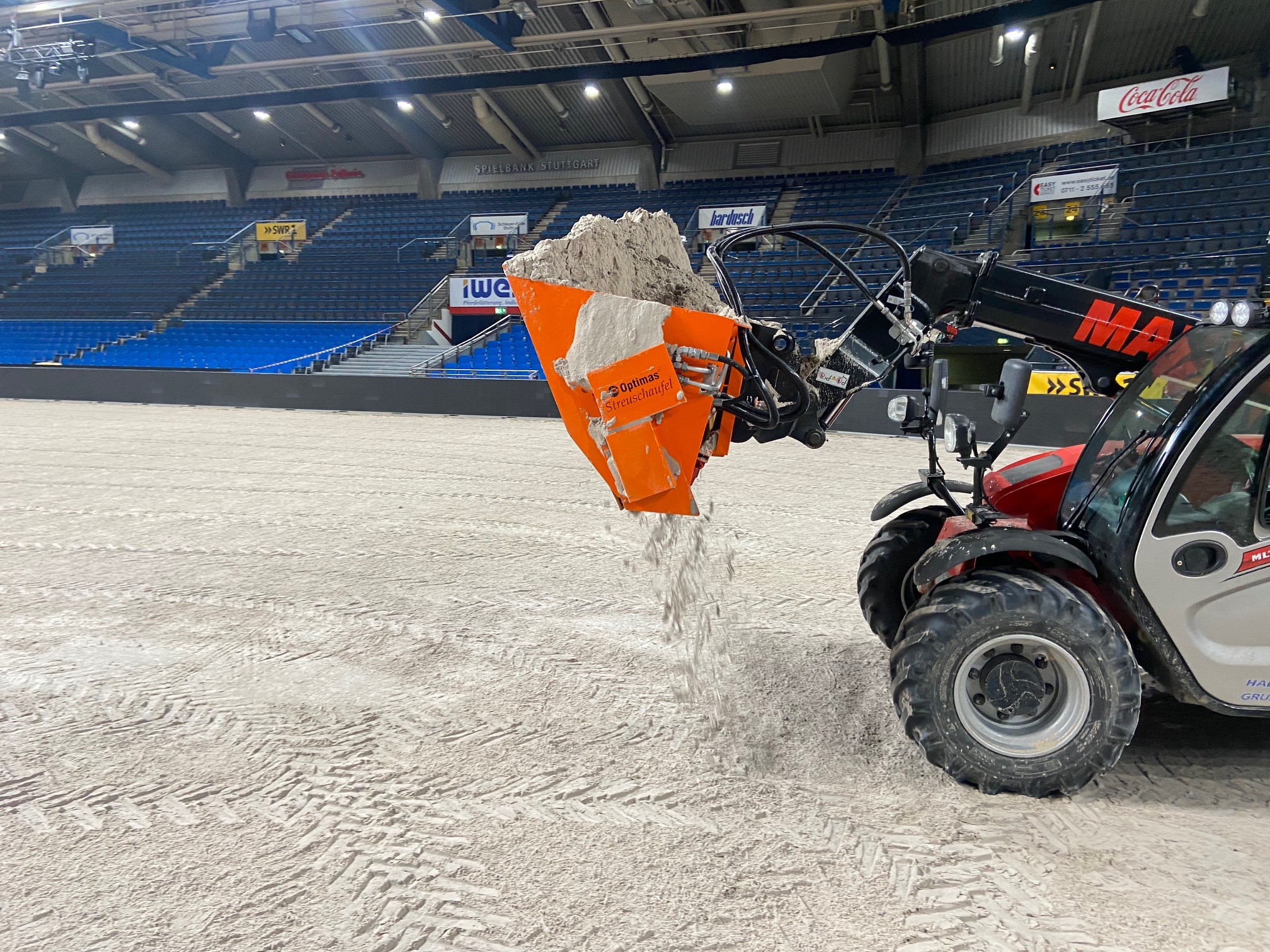 Rot-silberne landwirtschaftliche Maschine, die Sand auf einer Bühne in einem Stadion ebnet und sich mit Optimas-Geräten auf eine Veranstaltung vorbereitet – Optimas