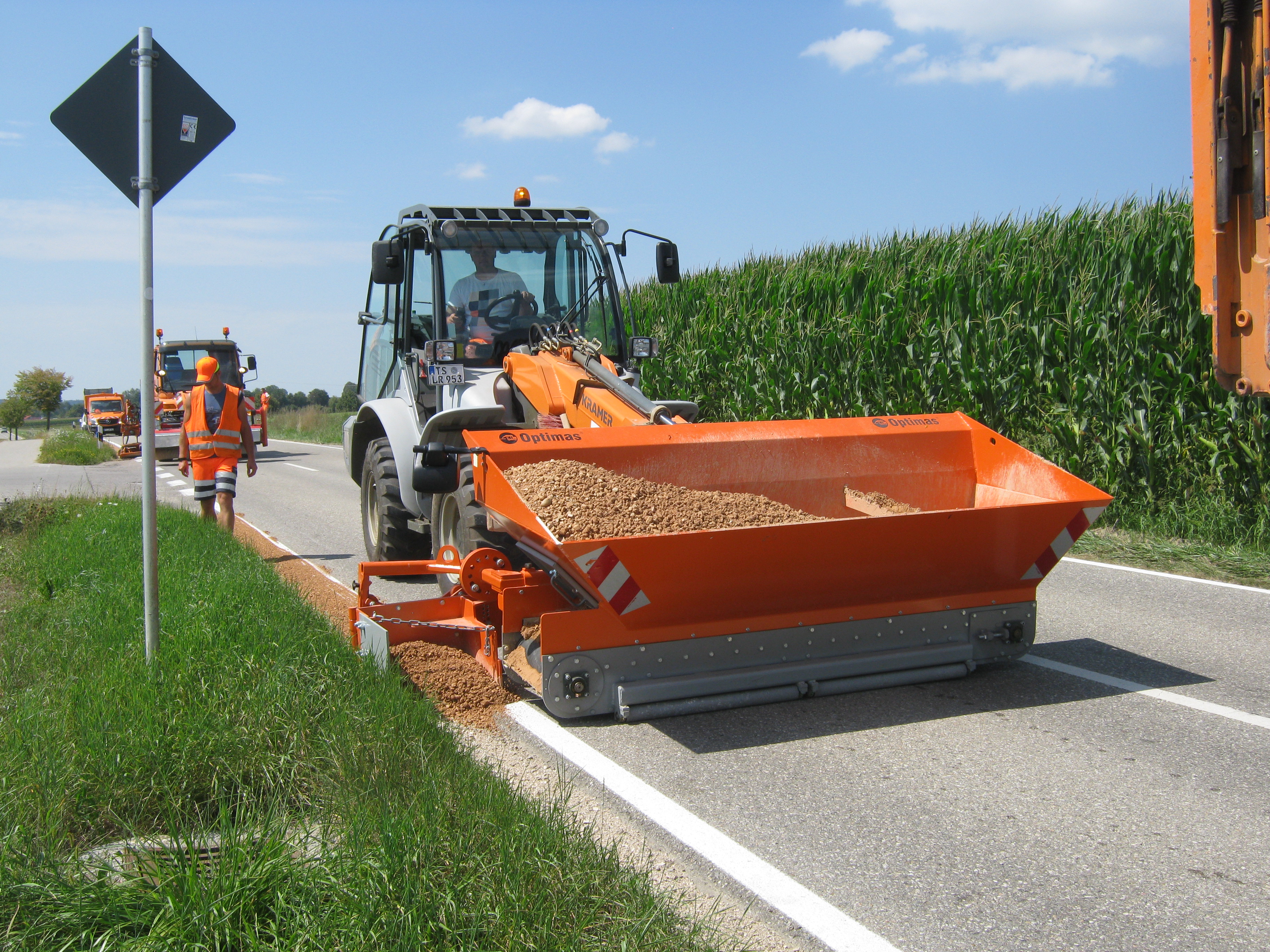 Straßeninstandhaltungsfahrzeug verteilt Kies am Straßenrand und demonstriert effiziente Instandhaltung der Infrastruktur, eine Optimas-Ausrüstung und blauen Himmel – Optimas.