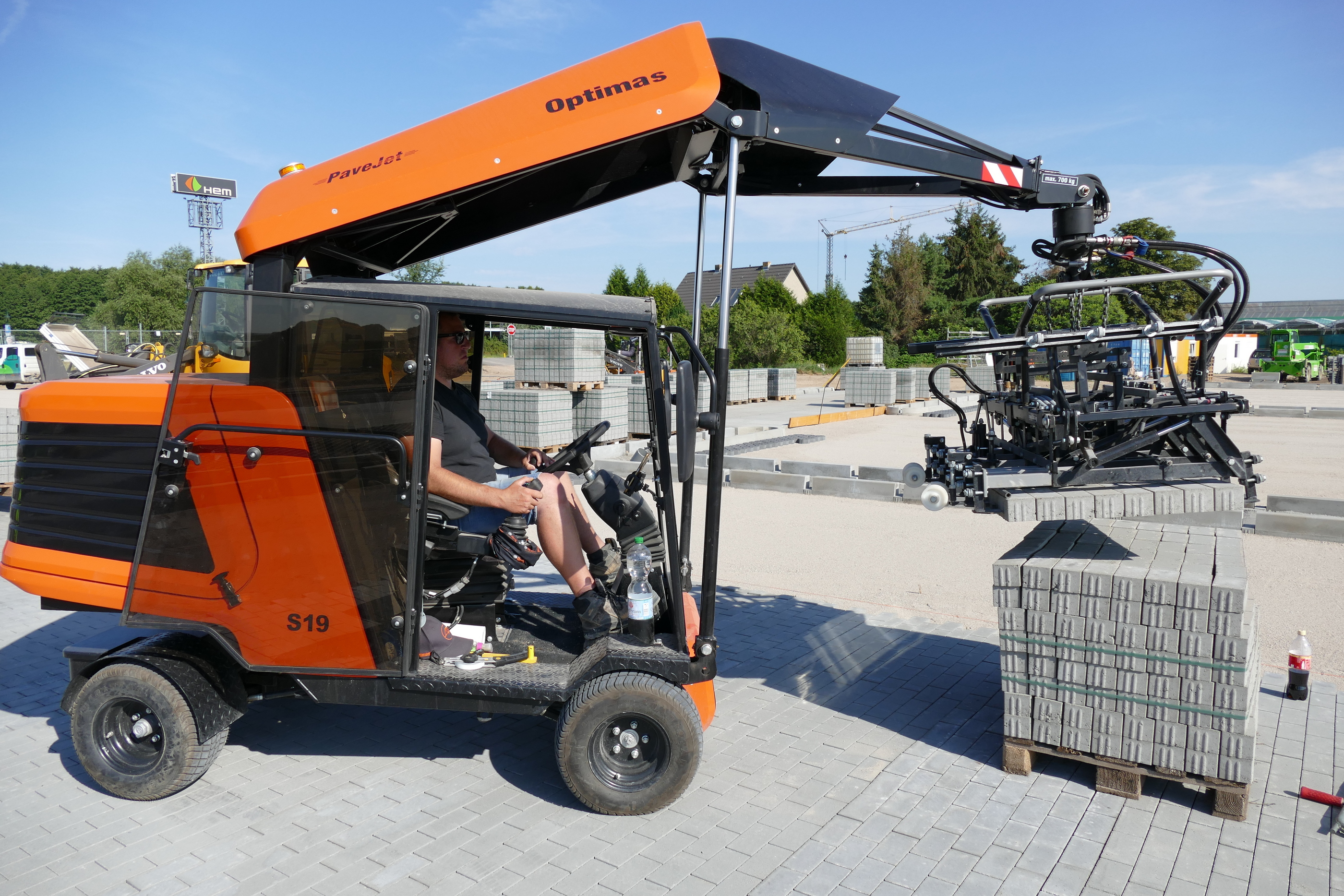 Pflastermaschine Optimas S19 im Einsatz, bedient von einem Arbeiter mit Sonnenbrille, der Pflastersteine ​​aufnimmt, mit sichtbaren Steinpaletten – Optimas
