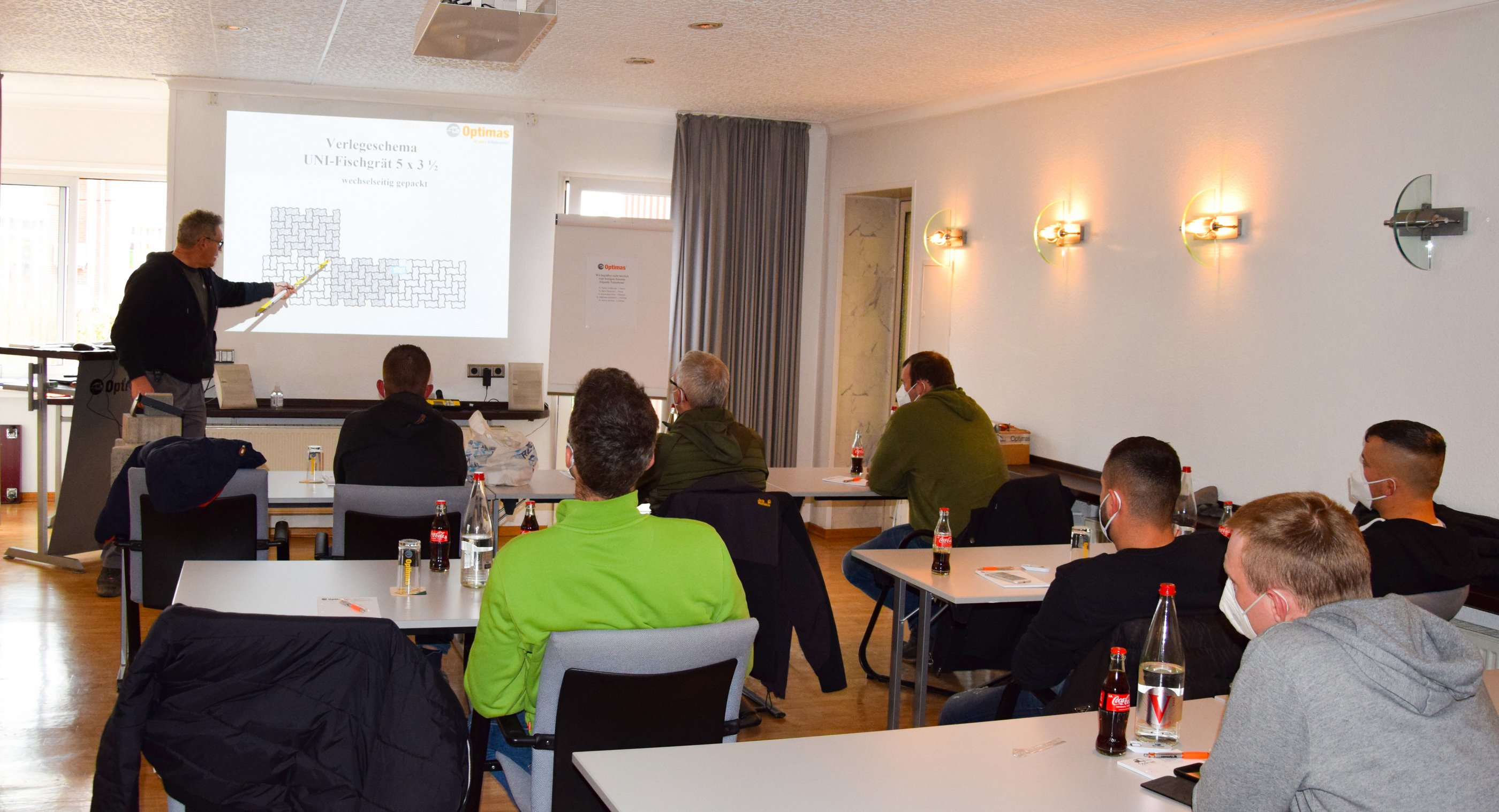 Teilnehmer eines Seminars zum Thema Pflastersteinmuster lauschen einer Präsentation, während sie an Tischen sitzen, einige tragen Gesichtsmasken – Optimas.