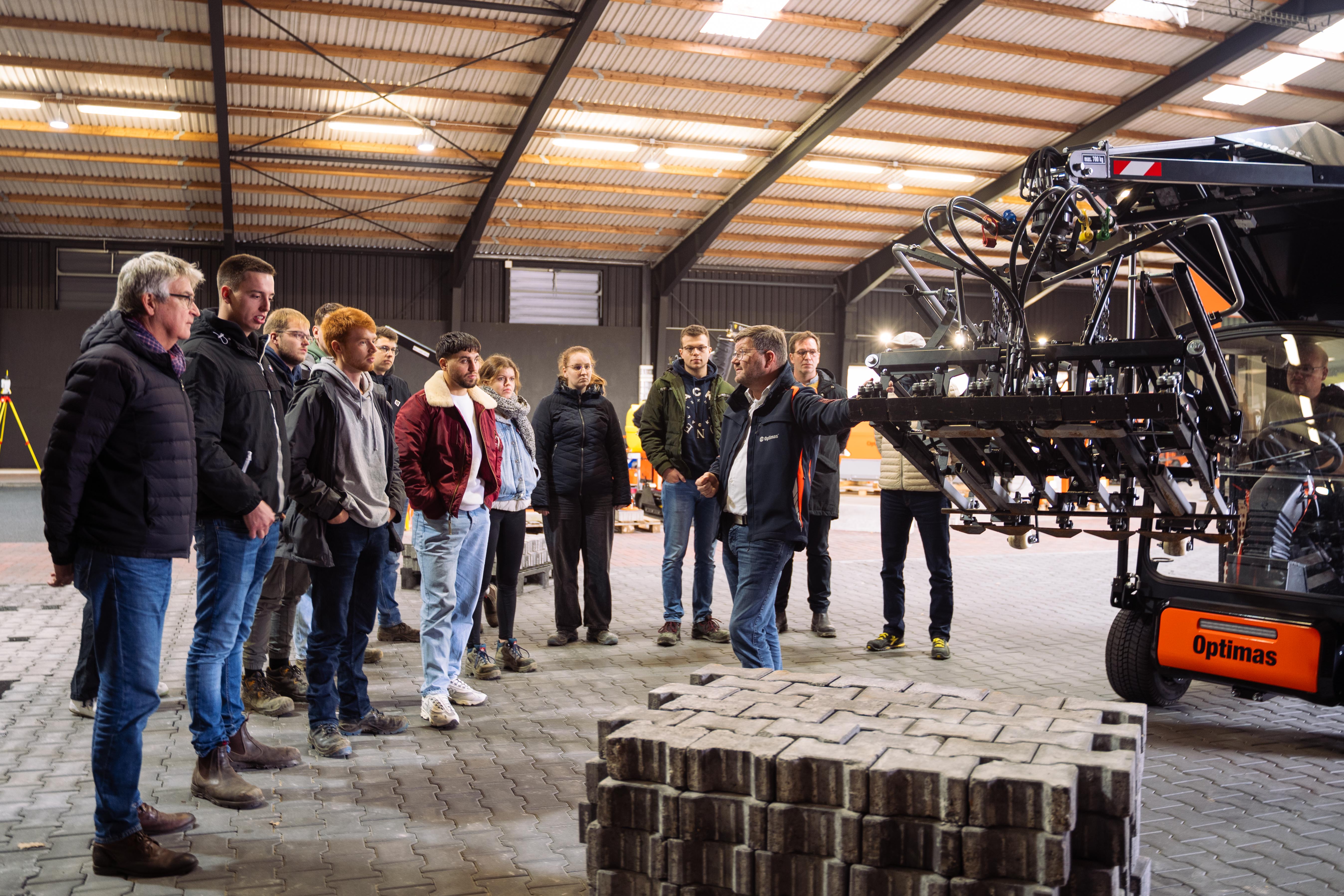 Eine Gruppe von Menschen beobachtet eine Maschinendemonstration in einem Lagerhaus und konzentriert sich dabei auf Gerätedetails – Optimas.