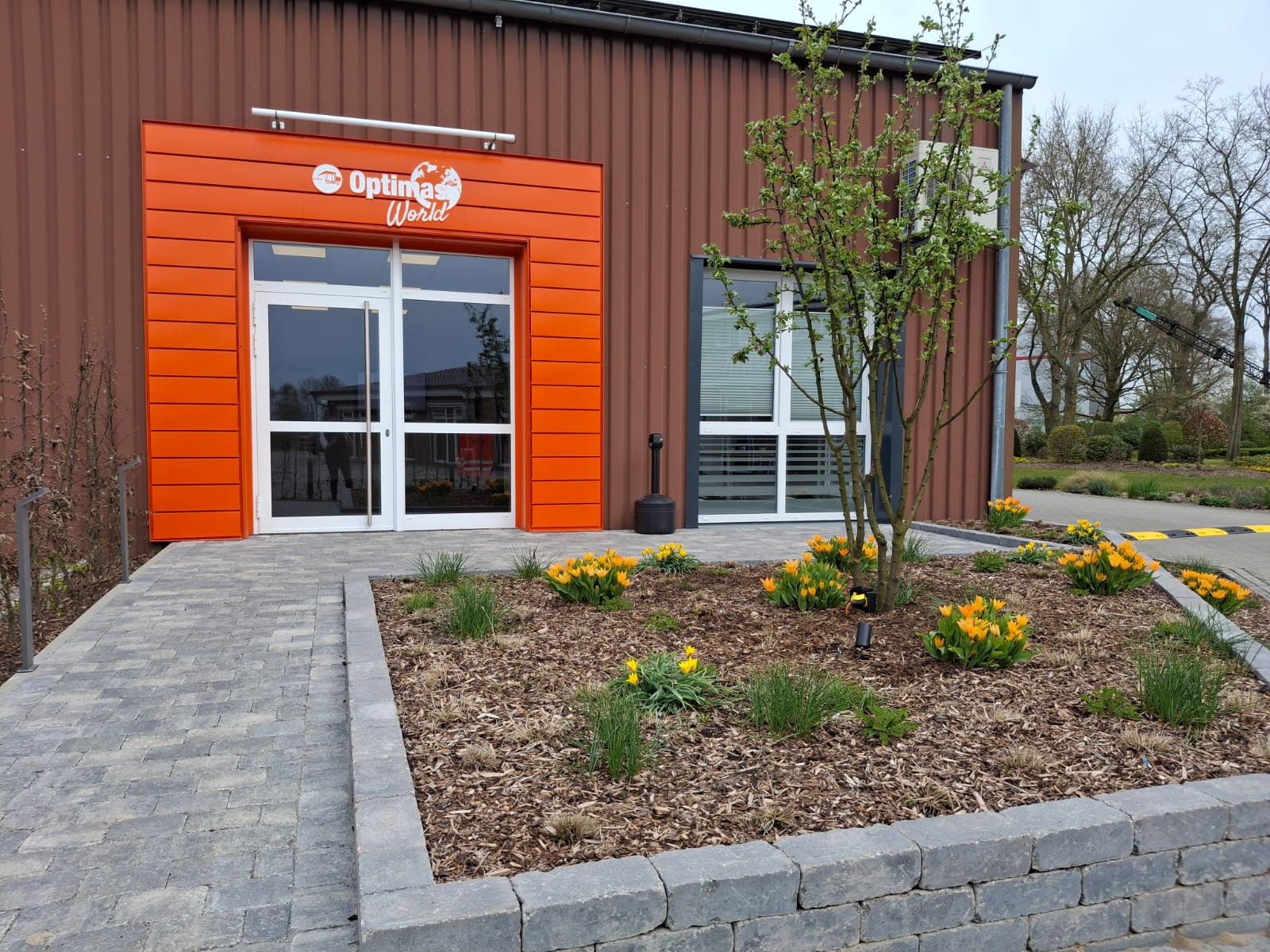 Eingang der Optimas-Welt mit leuchtend orangefarbener Fassade und angelegten Blumenbeeten – Optimas.