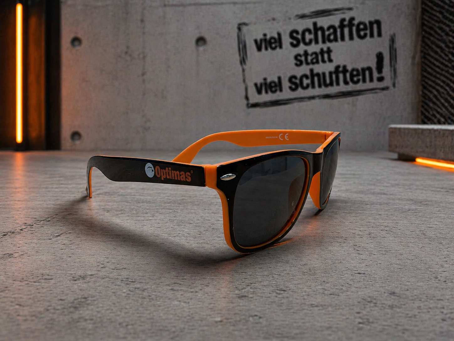 Stilvolle Sonnenbrille in Schwarz und Orange auf einer strukturierten Oberfläche mit einem motivierenden Zitat im Hintergrund – Optimas.