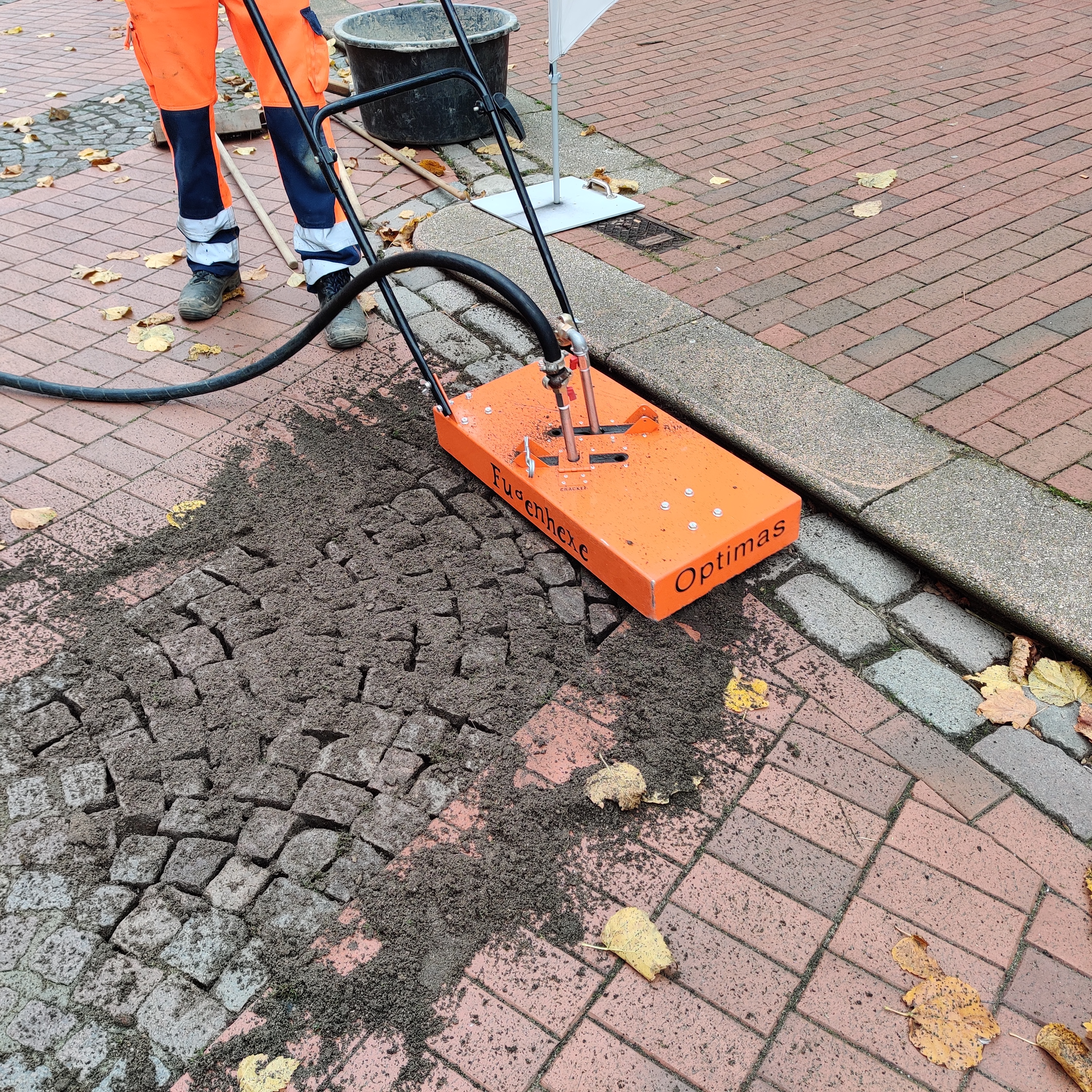 Ein Wartungsarbeiter verwendet eine orangefarbene Fugenreinigungsmaschine auf einer Ziegel- und Kopfsteinpflasteroberfläche mit der Aufschrift „Optimas“, um Zugänglichkeit und Sicherheit zu gewährleisten – Optimas.
