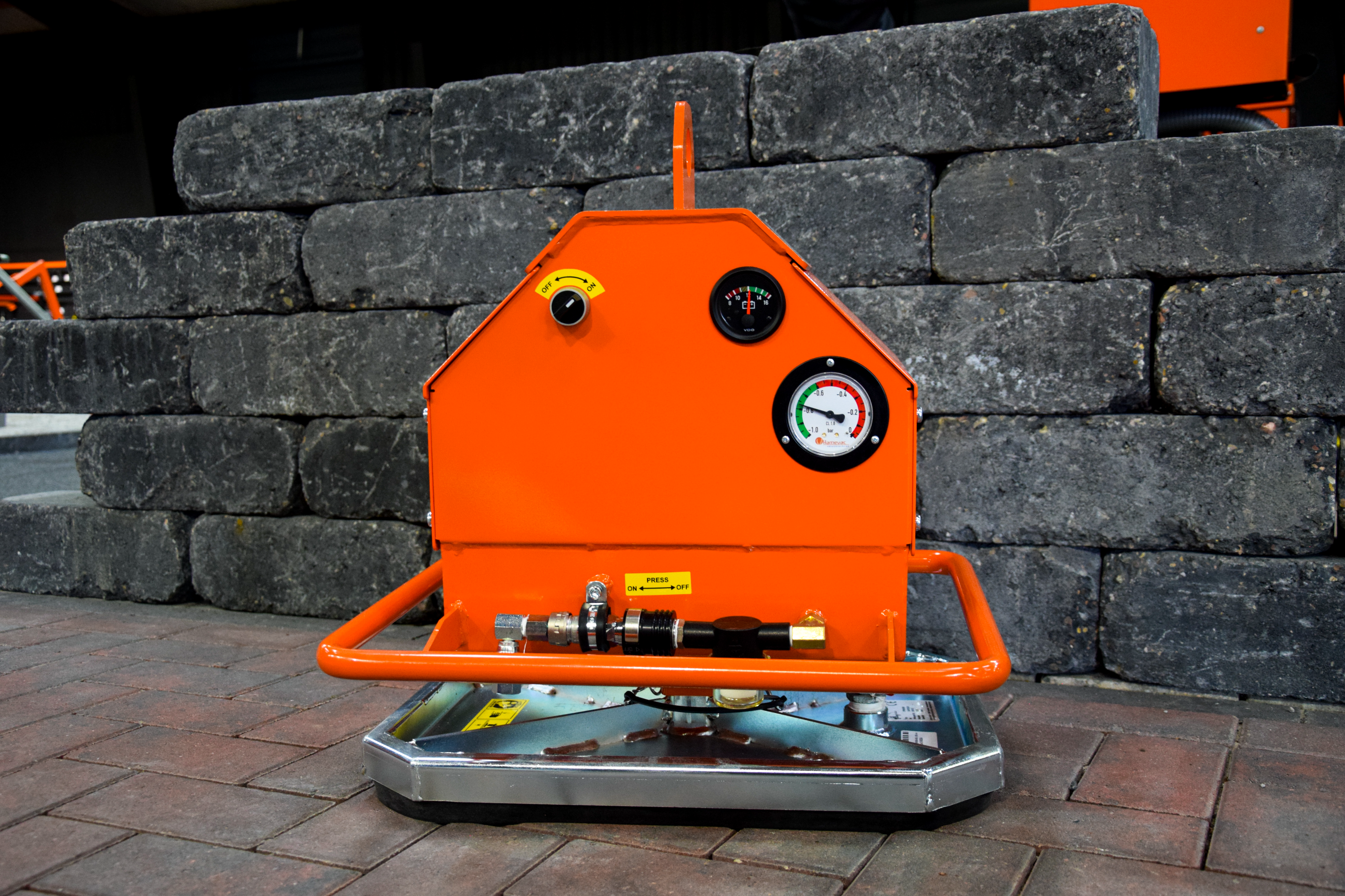 Ein orangefarbenes Vakuumhebegerät mit Messgeräten und Metallsockel steht auf einer gepflasterten Fläche vor Steinblöcken – Optimas.