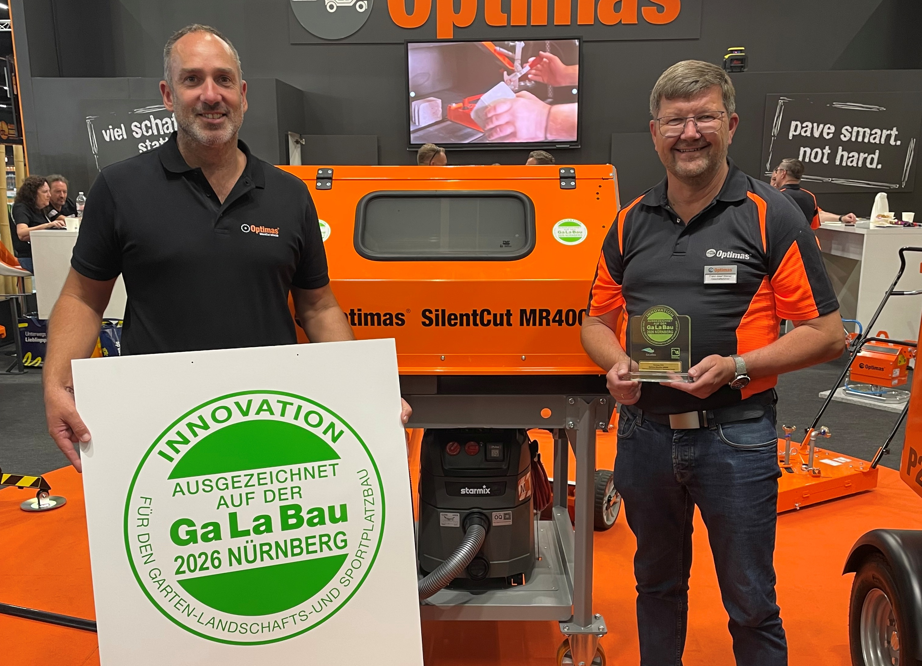Zwei Männer stehen vor einer orangefarbenen Optimas SilentCut MR400-Maschine und halten ein Innovationspreisschild – Optimas.
