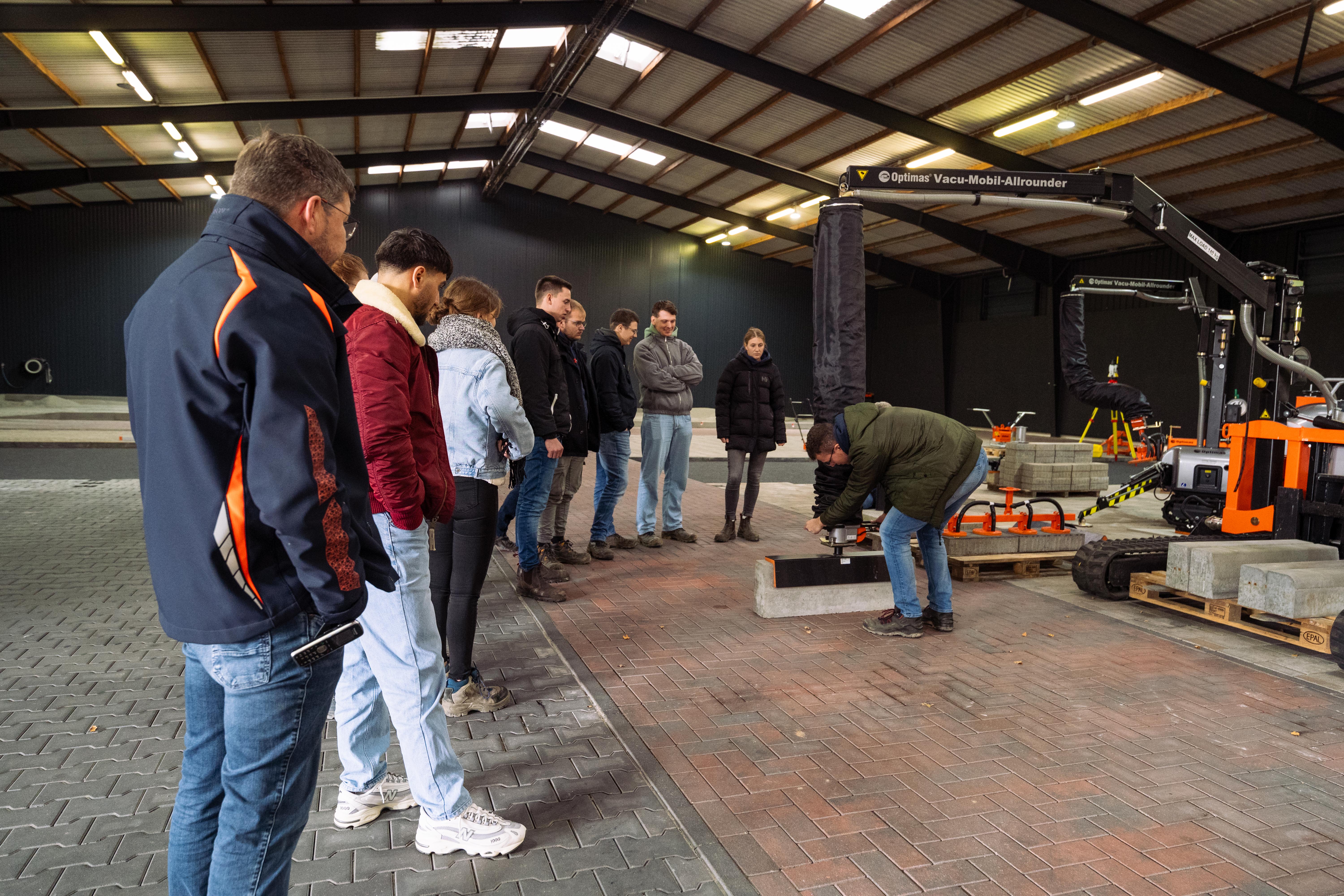 Eine Gruppe von Menschen beobachtet die Vorführung eines Bauwerkzeugs in einer weitläufigen Industrieanlage – Optimas.