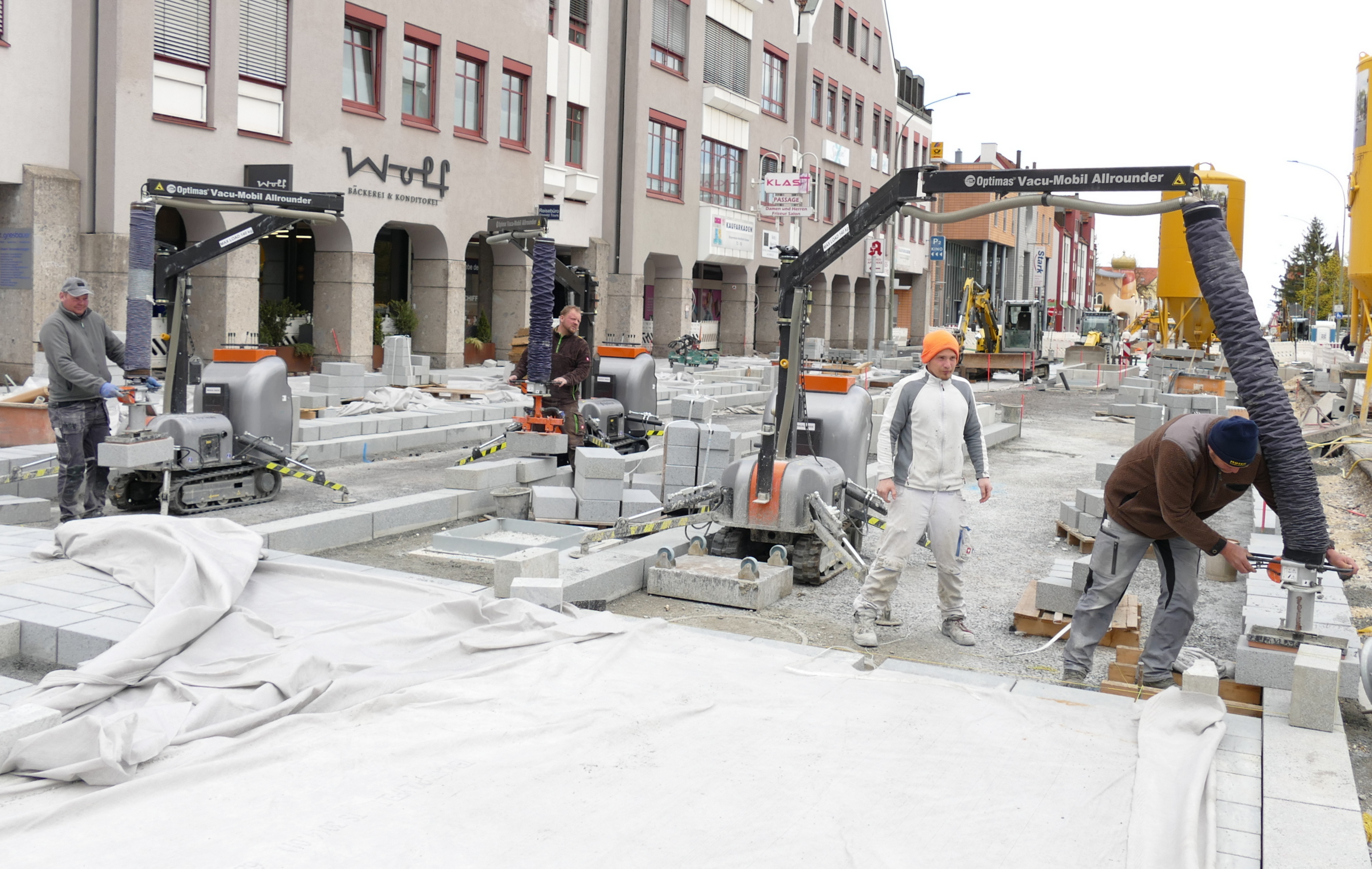 Straßenbau im Gange, wobei Arbeiter mit Optimas Vacu-Mobil Allrounder-Maschinen Pflastersteine ​​verlegen und eine europäische Stadtkulisse zeigen – Optimas.