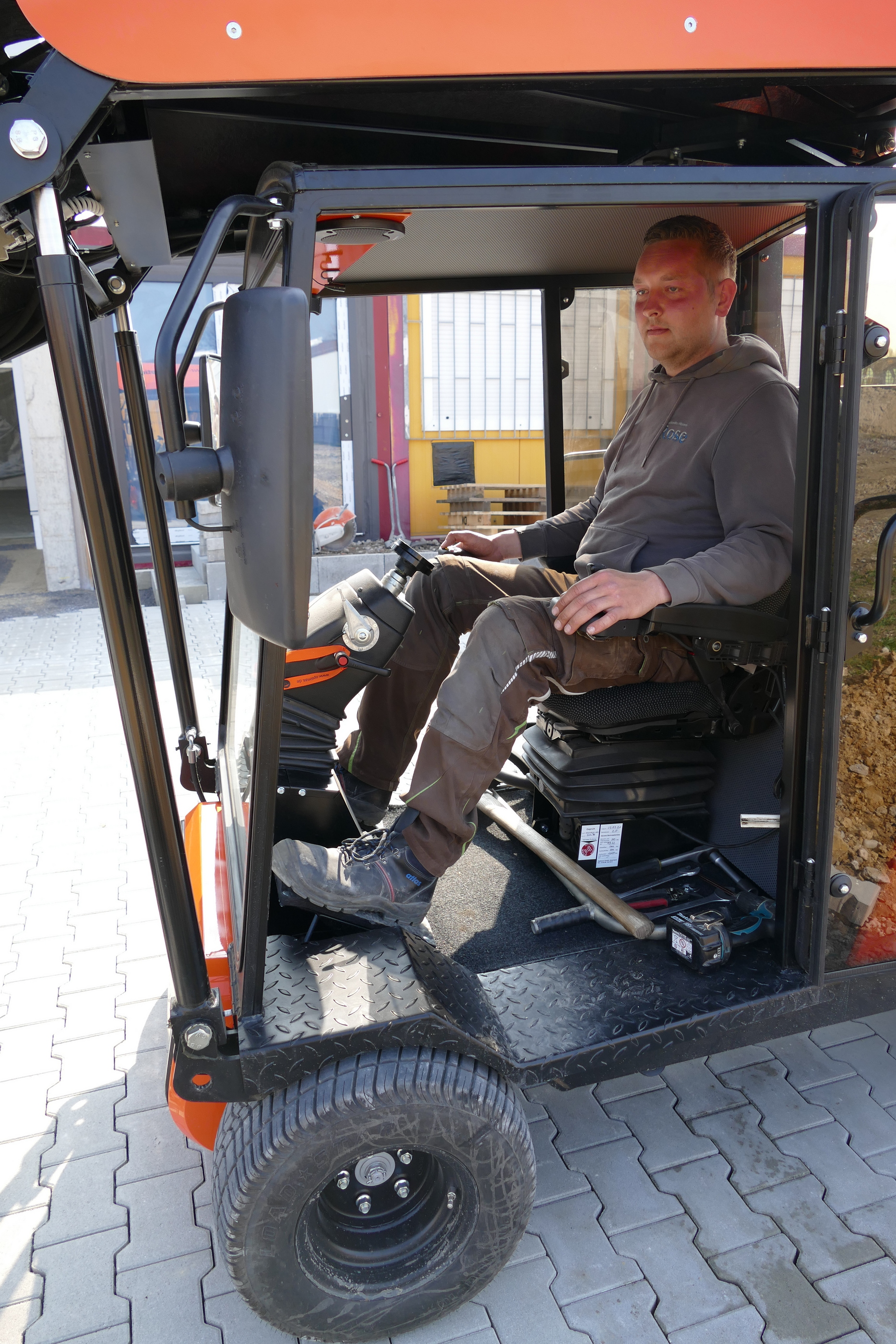 Mann in Arbeitskleidung bedient ein orange-schwarzes elektrisches Kompakt-Nutzfahrzeug und demonstriert Benutzerfreundlichkeit und moderne Funktionen – Optimas.