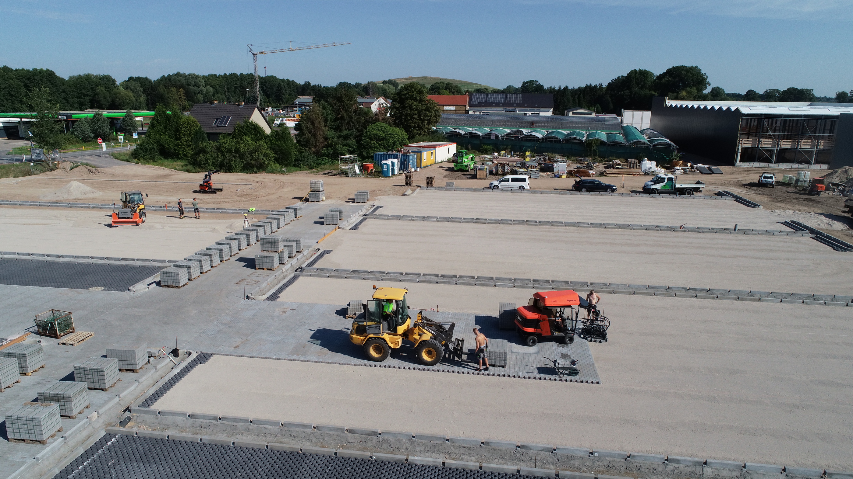 Baustelle mit Pflastersteinverlegung unter Einsatz von Maschinen und Arbeitern, darunter Bagger und Pflastermaschine. Betonpflastersteine ​​werden ordentlich gestapelt – Optimas.