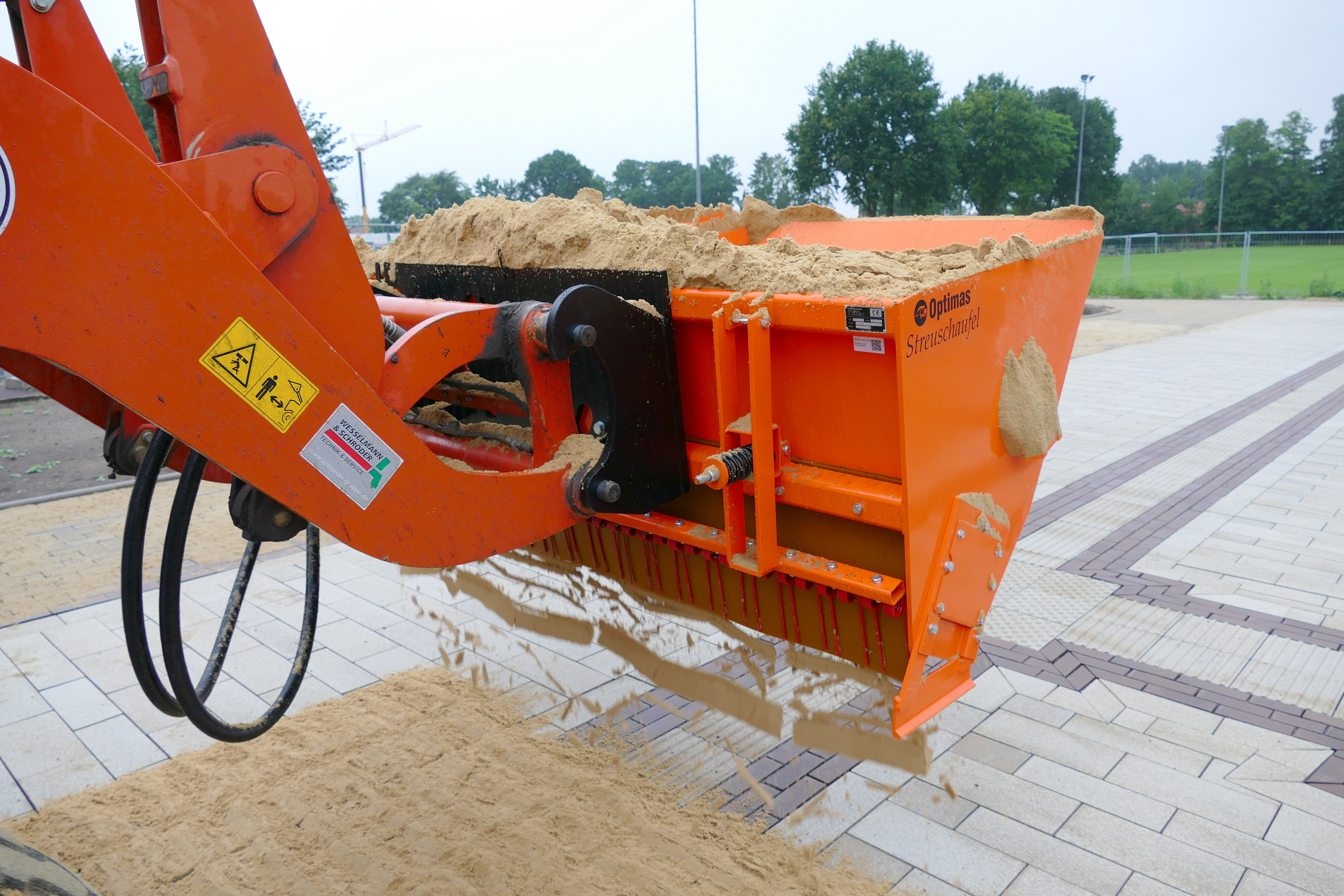 Nahaufnahme einer orangefarbenen Optimas-Streuschaufel, die mit Sand gefüllt ist und sich auf einer gepflasterten Oberfläche verteilt, wobei die Ausrüstung effizient arbeitet – Optimas.