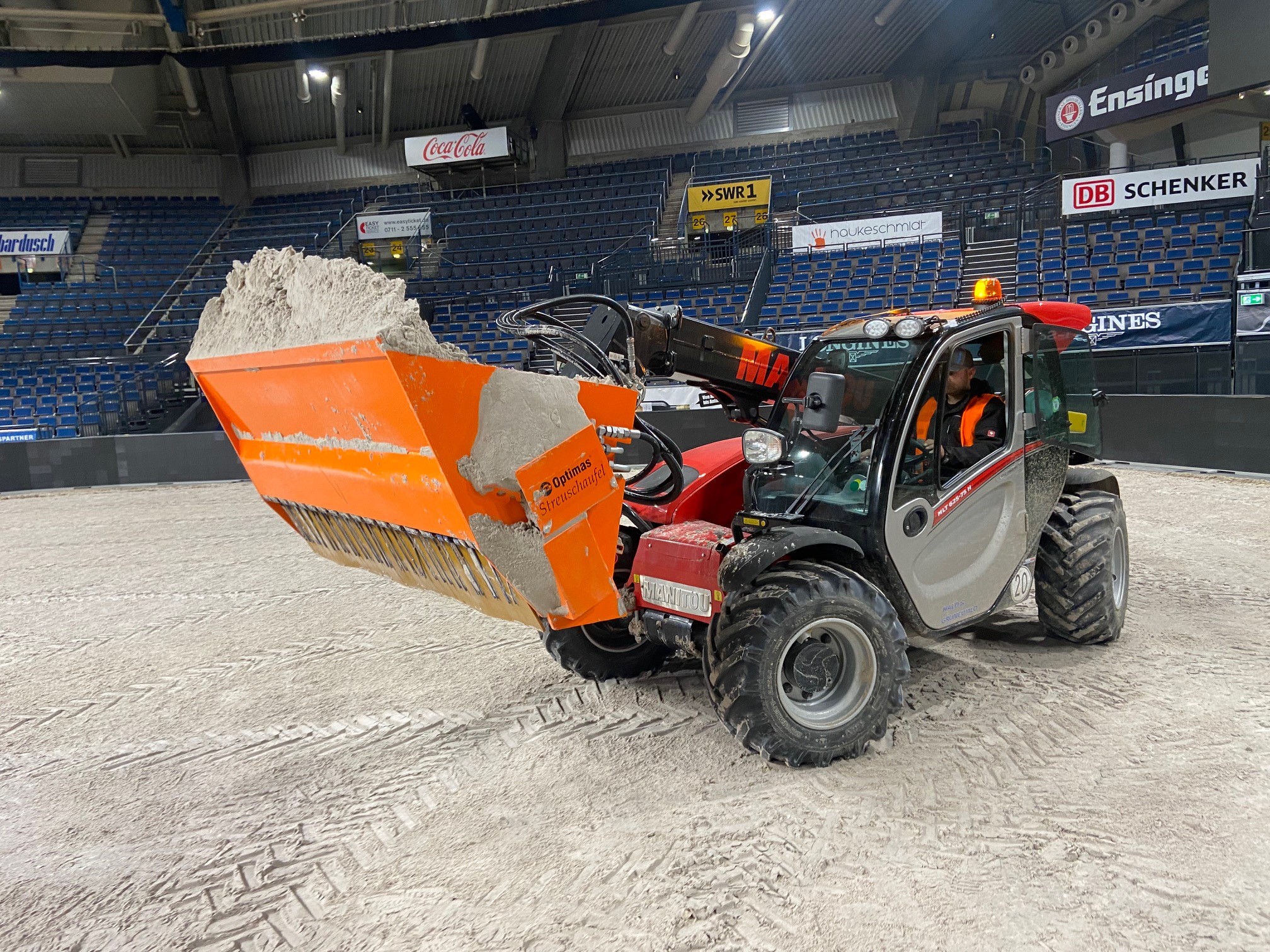 Der Frontlader ist mit einer orangefarbenen, mit weißem Material gefüllten Streuschaufel ausgestattet und reinigt den Arenaboden in einer Innenumgebung – Optimas