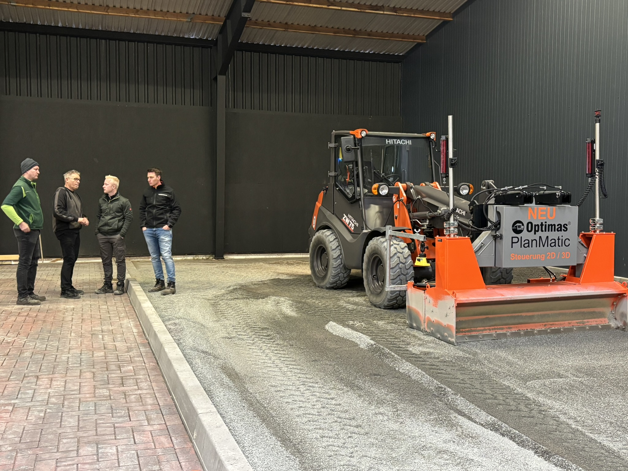 Vier Männer diskutieren in der Nähe einer Hitachi-Baumaschine mit Optimas PlanMatic-Technologie in einer Lagerhalle – Optimas.