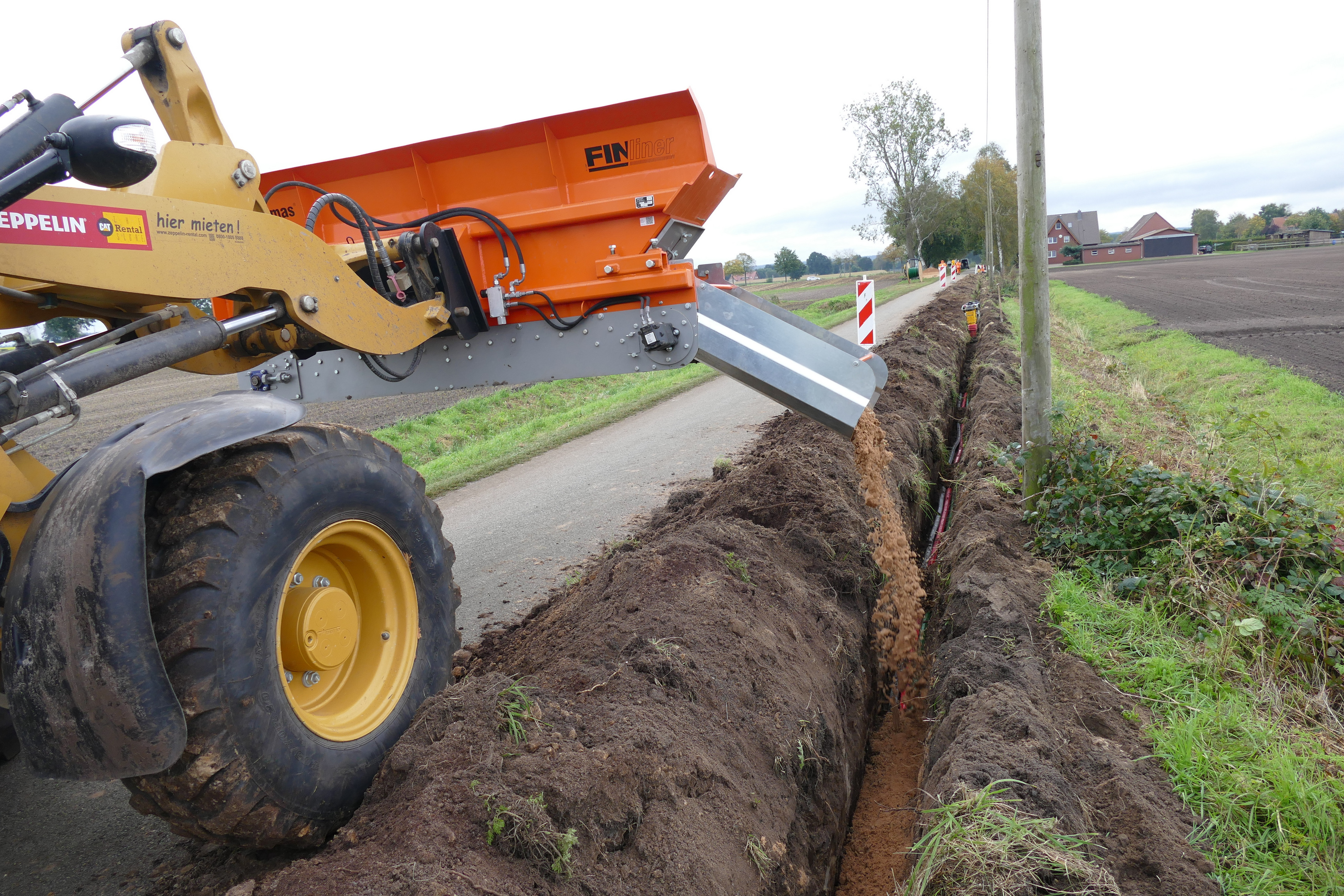 Ein gelber Bagger, der entlang einer Landstraße einen Graben mit Erde auffüllt, der zum Vergraben von Elektrokabeln dient, und Baumaschinen präsentiert – Optimas.
