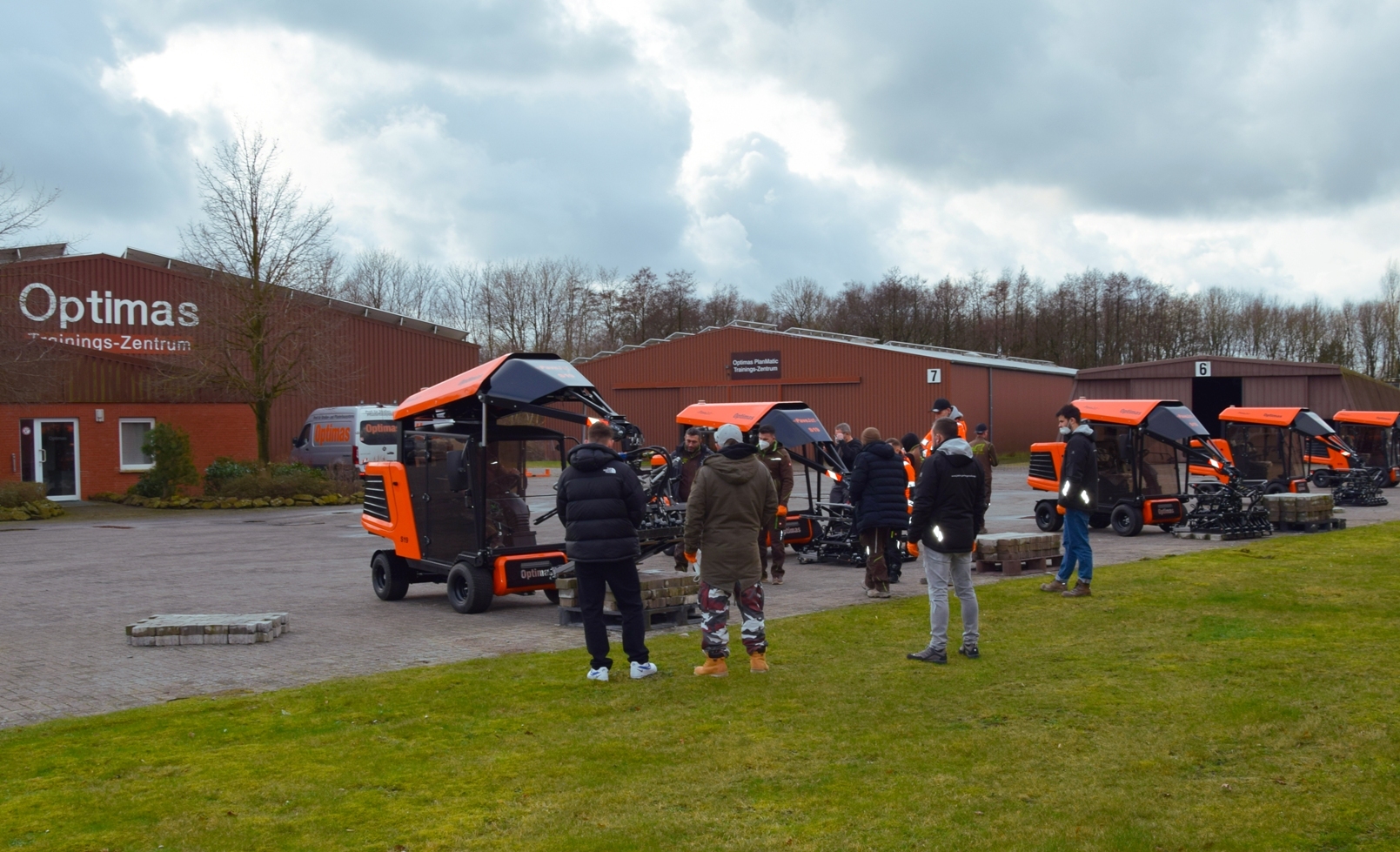 Außenaufnahme, die eine Gruppe von Menschen zeigt, die sich um orangefarbene Optimas PlanMatic-Maschinen im Optimas Training Center versammelt haben, bewölkter Himmel – Optimas.