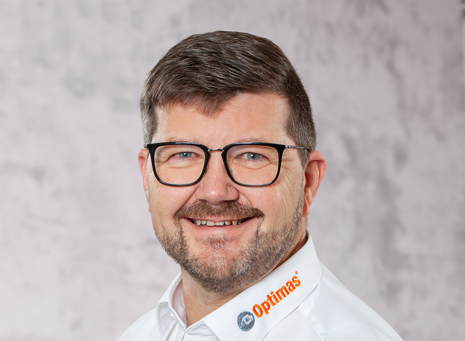 Ein lächelnder Mann mit Brille und Bart, der ein weißes Hemd mit dem Optimas-Logo – Optimas – trägt.