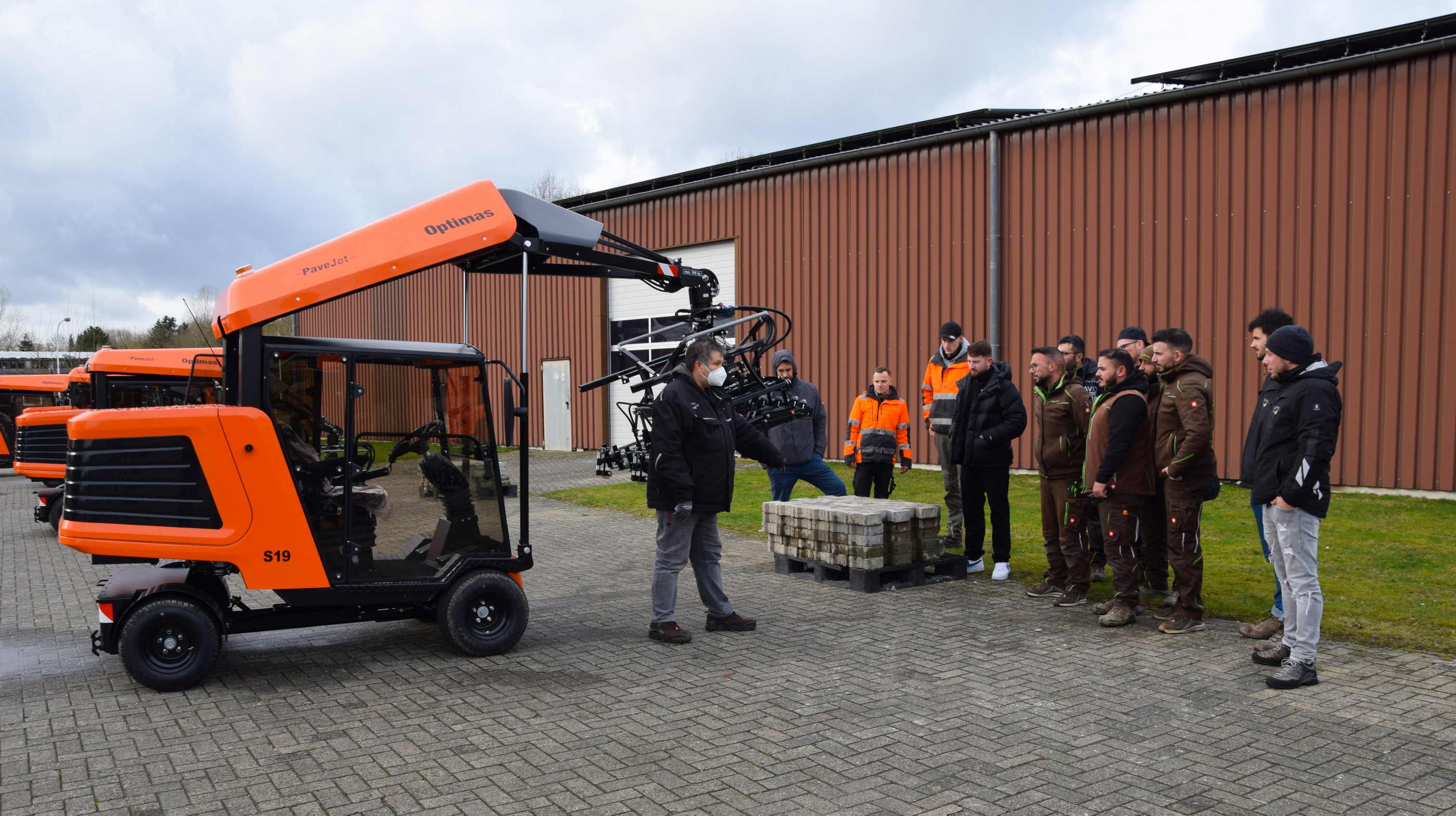 Gruppe von Menschen, die einer Erklärung zur Funktionsweise der orangefarbenen Optimas-Fertigerverlegemaschine zuhören, vor Ort – Optimas.