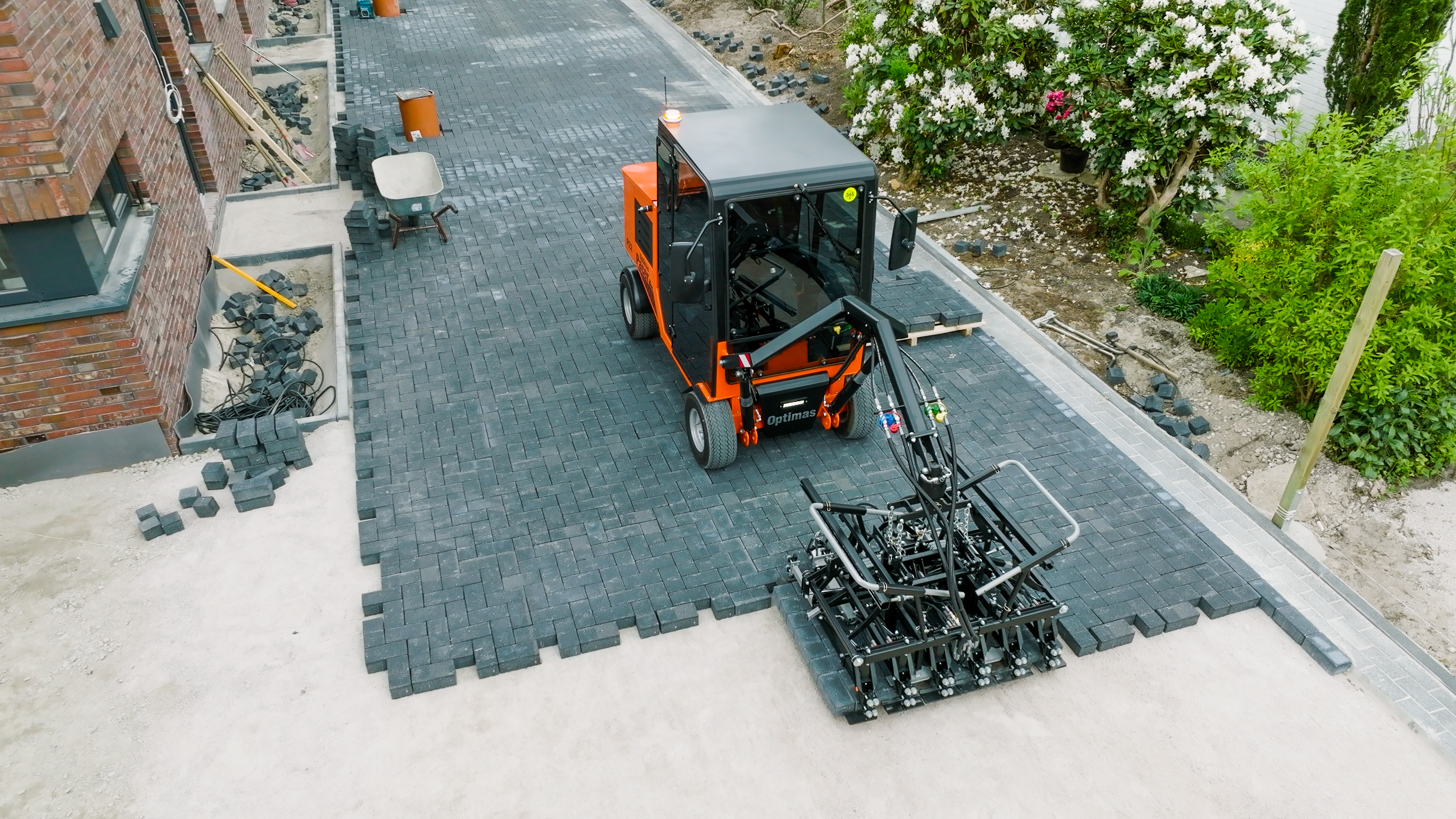 Draufsicht auf eine orangefarbene Optimas-Pflasterverlegemaschine, die sorgfältig graue Pflastersteine ​​auf einer Einfahrtsbaustelle verlegt – Optimas.