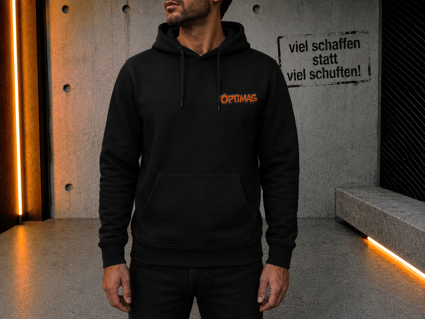 Ein Mann, der einen schwarzen Kapuzenpullover mit dem Aufdruck „Optimas“ trägt und vor einer strukturierten Wand steht – Optimas.