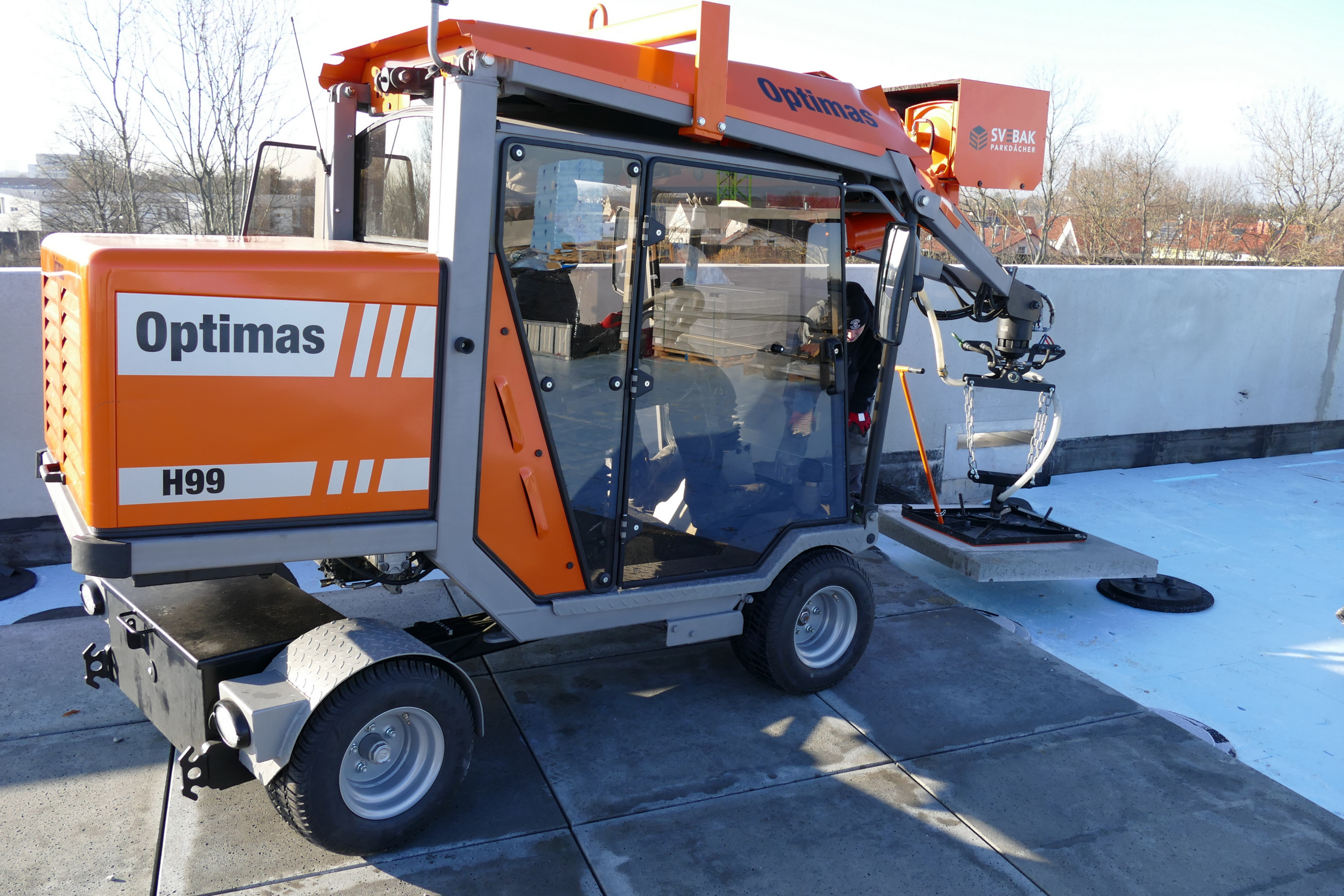 Verlegemaschine Optimas H99, grau und orange, mit Glaskabine, Arbeiten auf einem Dach mit Vakuumaufsatz zum Transport von Betonpflastersteinen – Optimas.