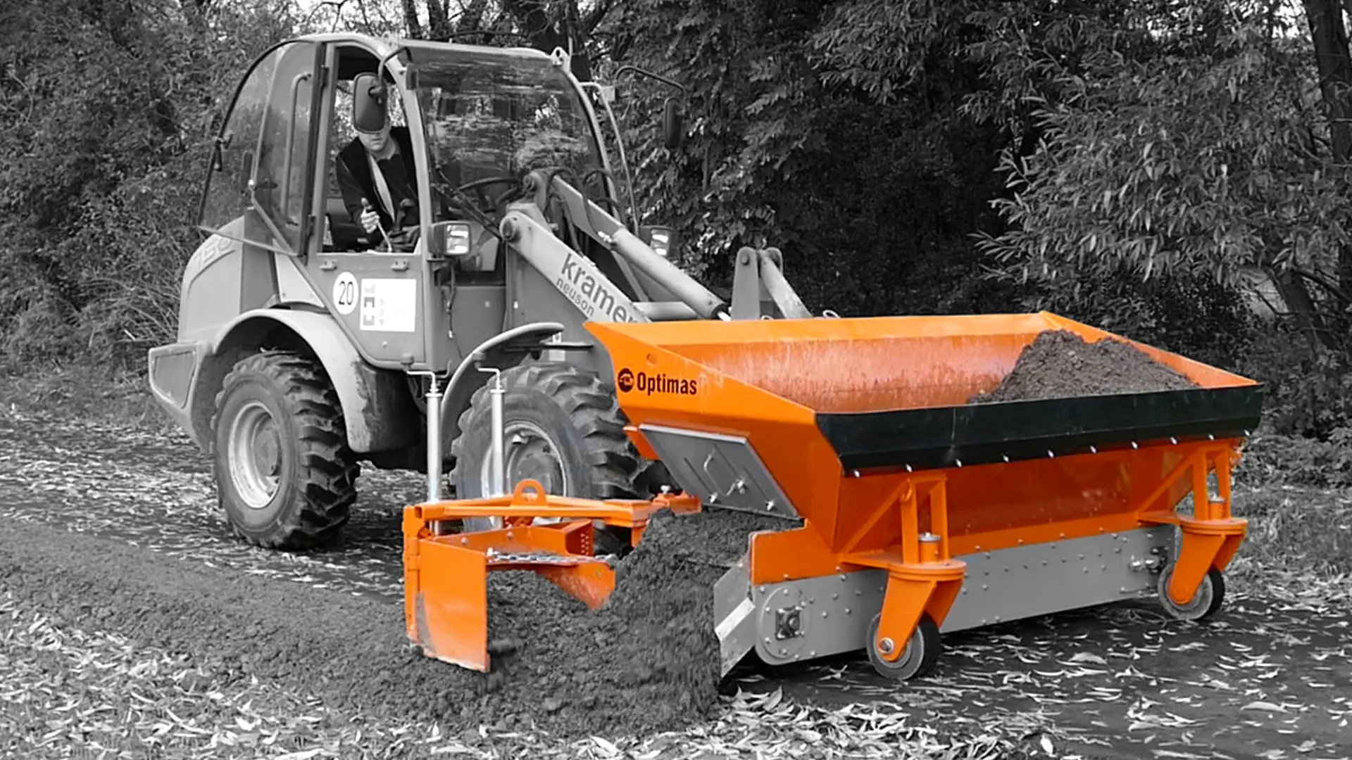 Ein Baufahrzeug fährt auf einem Feldweg und verteilt mit einem orangefarbenen Anbaugerät Erde – Optimas.
