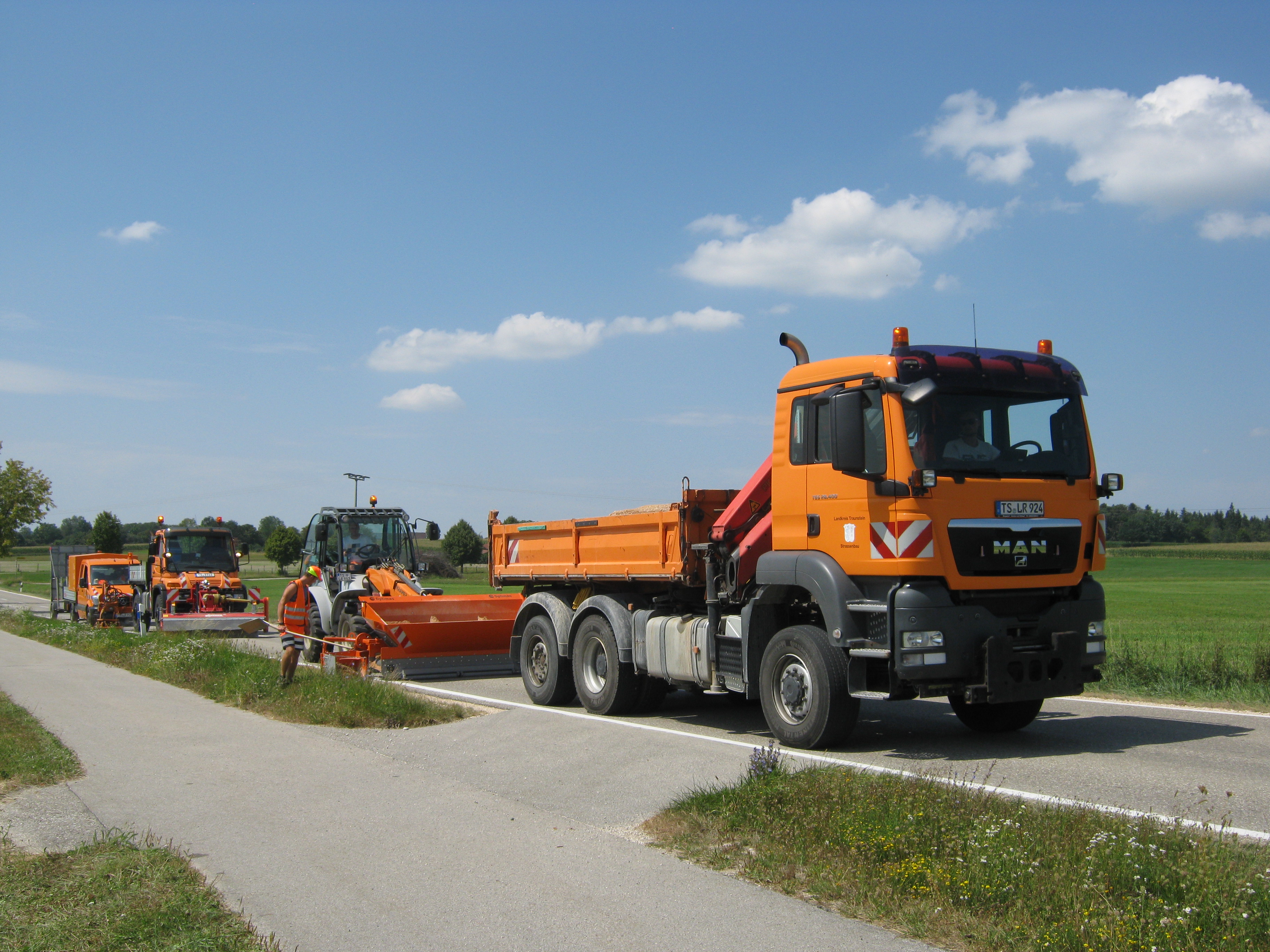 Ein leuchtend orangefarbener MAN-Lkw, gefolgt von Straßenmeistereifahrzeugen, fährt unter strahlend blauem Himmel über eine graue Asphaltstraße und sorgt für Verkehrssicherheit – Optimas.