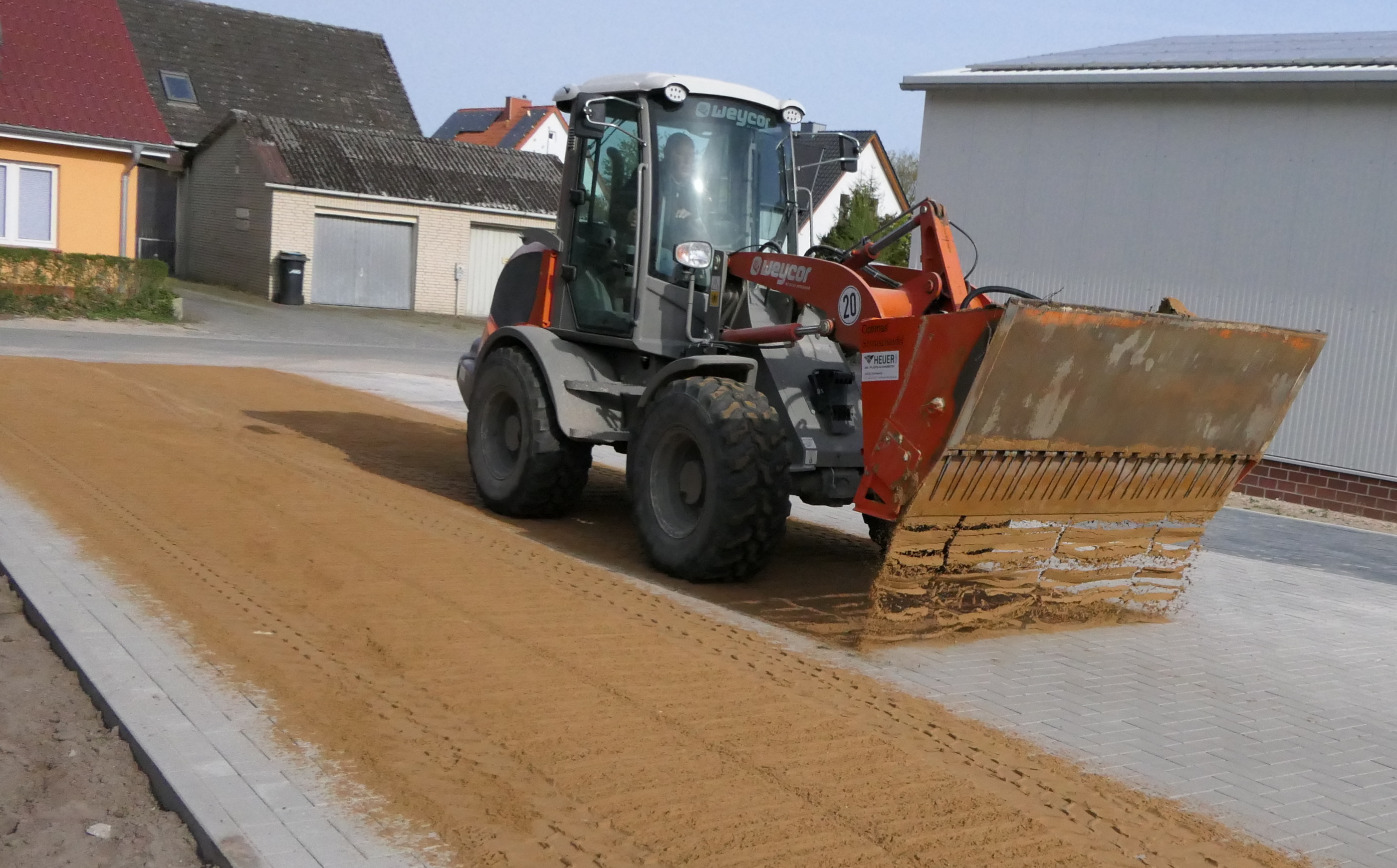 Ein Baufahrzeug mit Frontlader verteilt Sand auf einer neu asphaltierten Straße in einem Wohngebiet – Optimas.